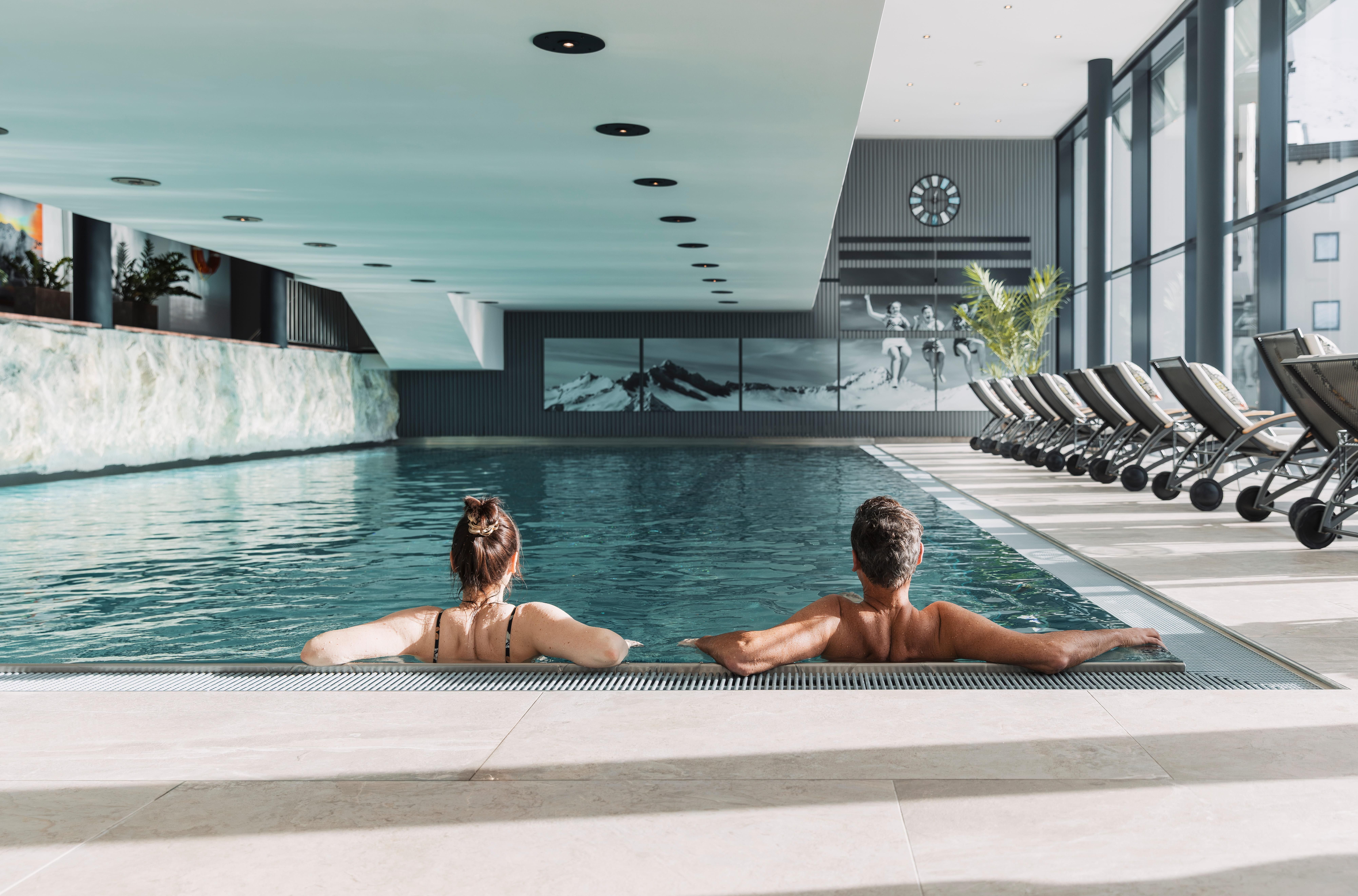 A modern indoor pool with relaxing loungers and large windows. Two people enjoy the tranquility at the poolside.