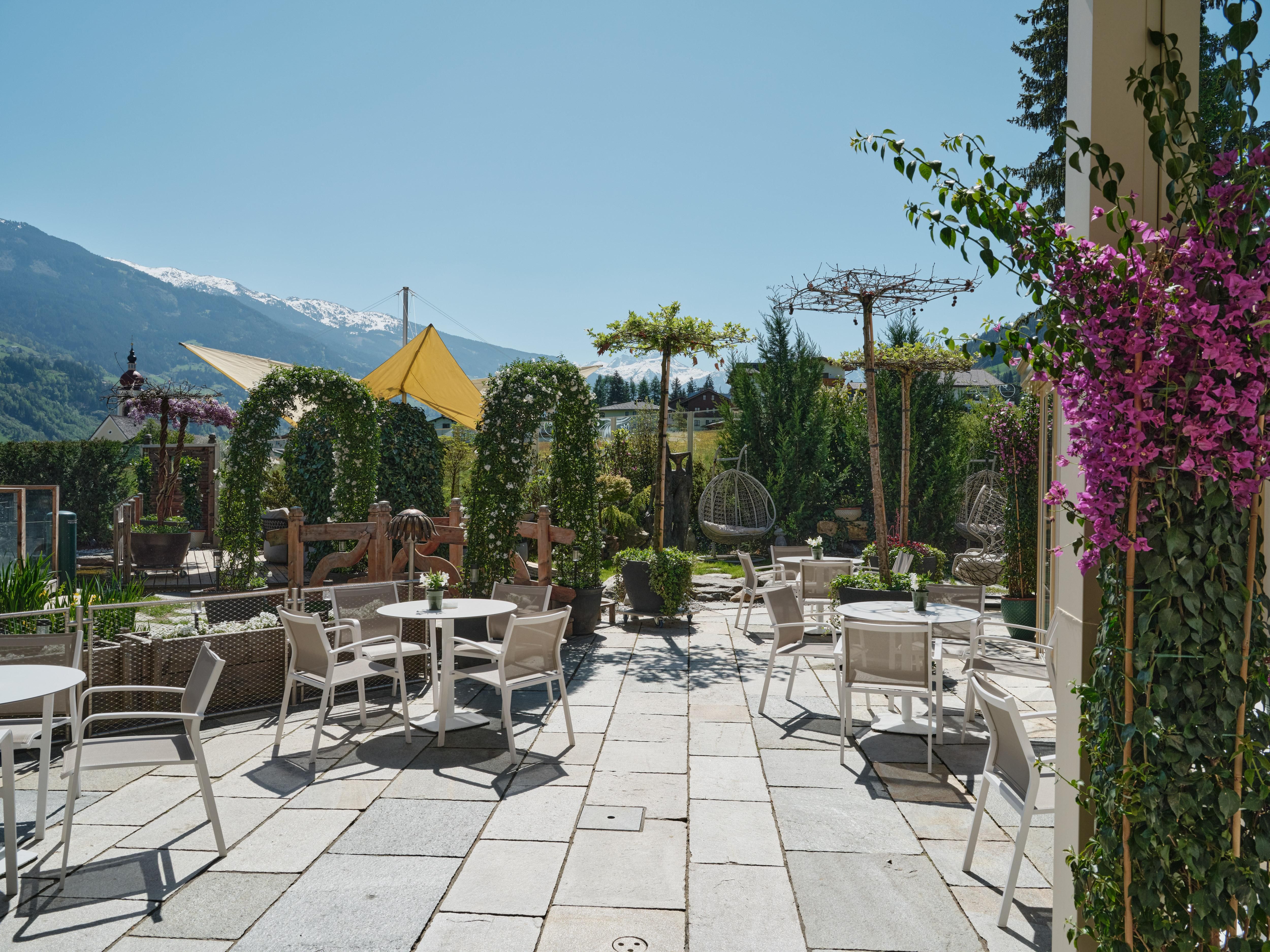 A beautiful outdoor area with tables and chairs, surrounded by plants and mountains in the background. The atmosphere is relaxed and inviting.