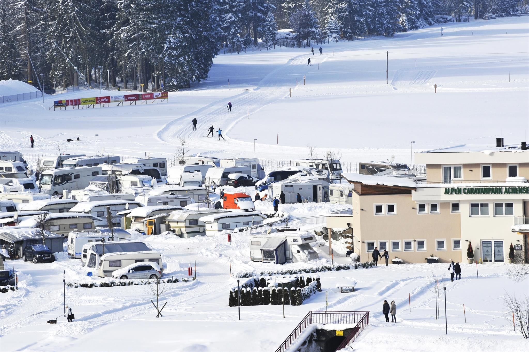 A snow landscape with motorhomes and a building in the foreground. In the background, cross-country skiers are visible on a ski trail.