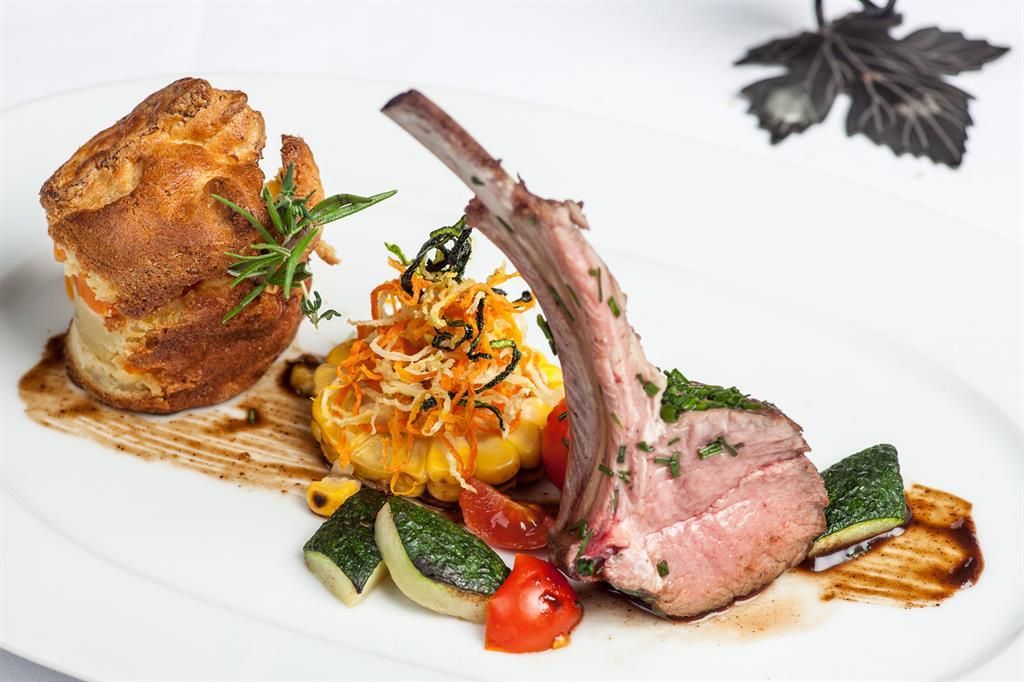 An elegant dish with lamb chops, colorful vegetables, and a side of corn. The dish is artfully presented and garnished with fresh herbs.
