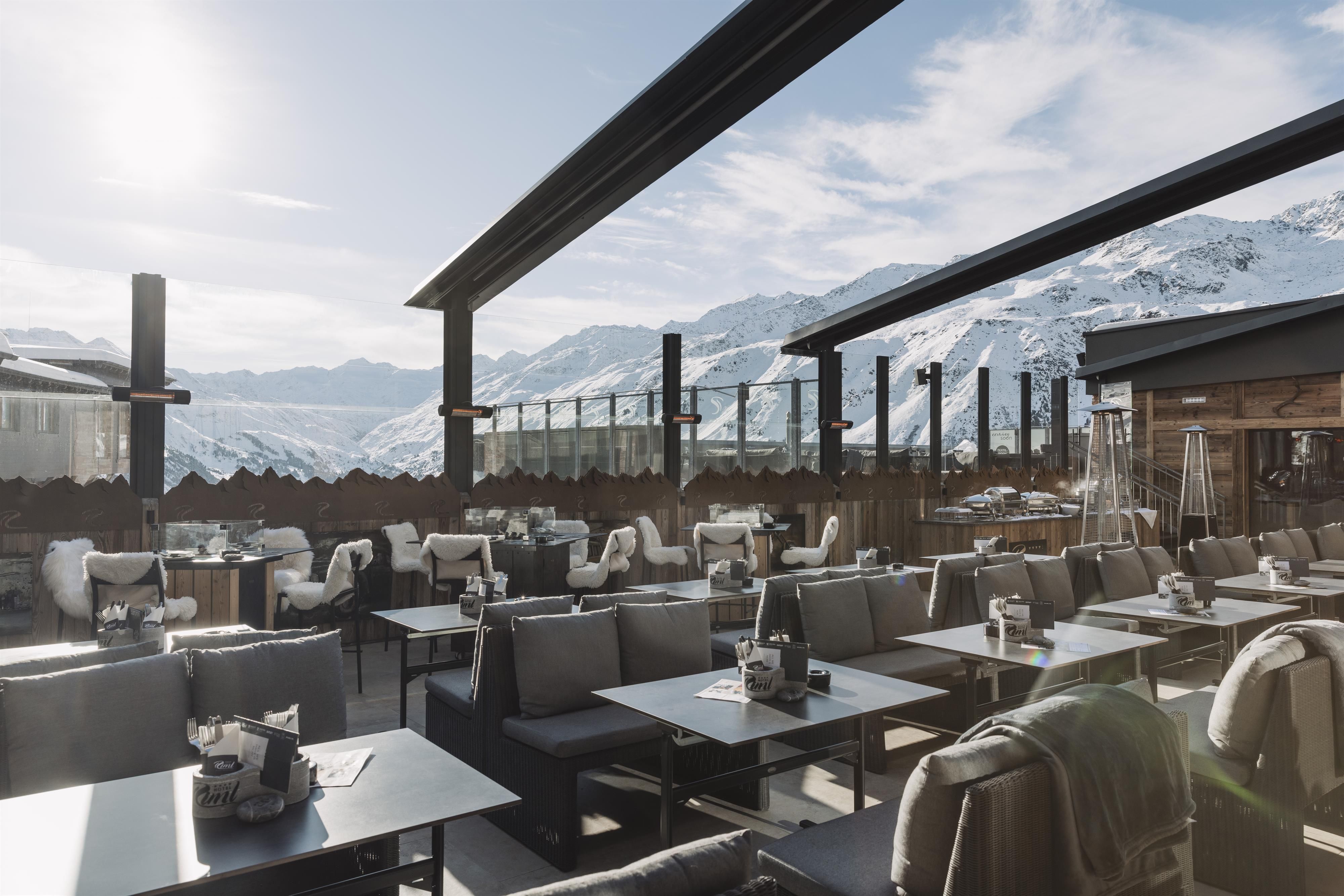A modern terrace of a restaurant overlooking snow-covered mountains. The tables are elegantly set and the sun is shining on the cozy seating.