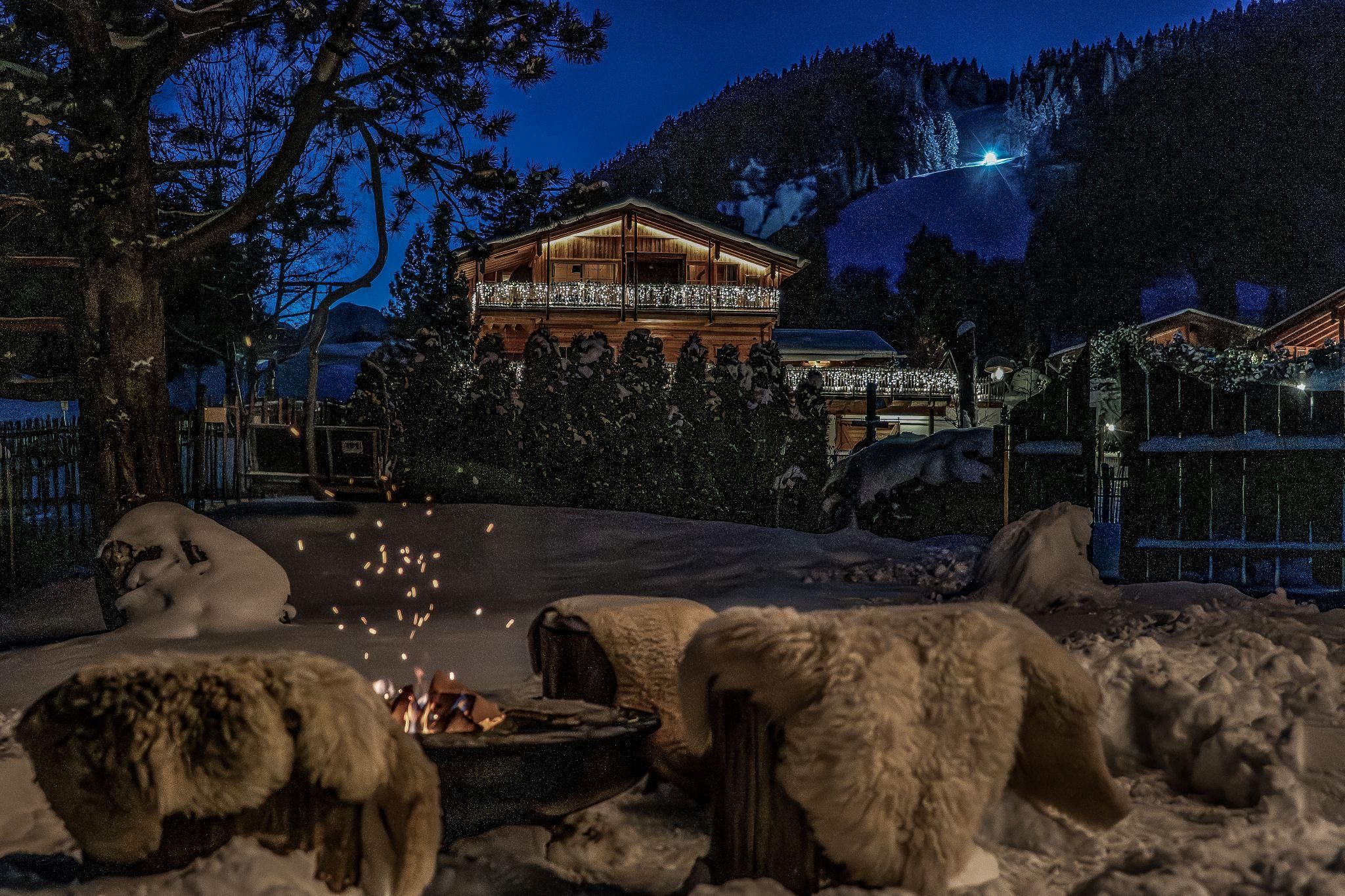 A cozy garden in the snow with a fire pit. In the background, there is a beautiful chalet and a wintry mountain landscape.