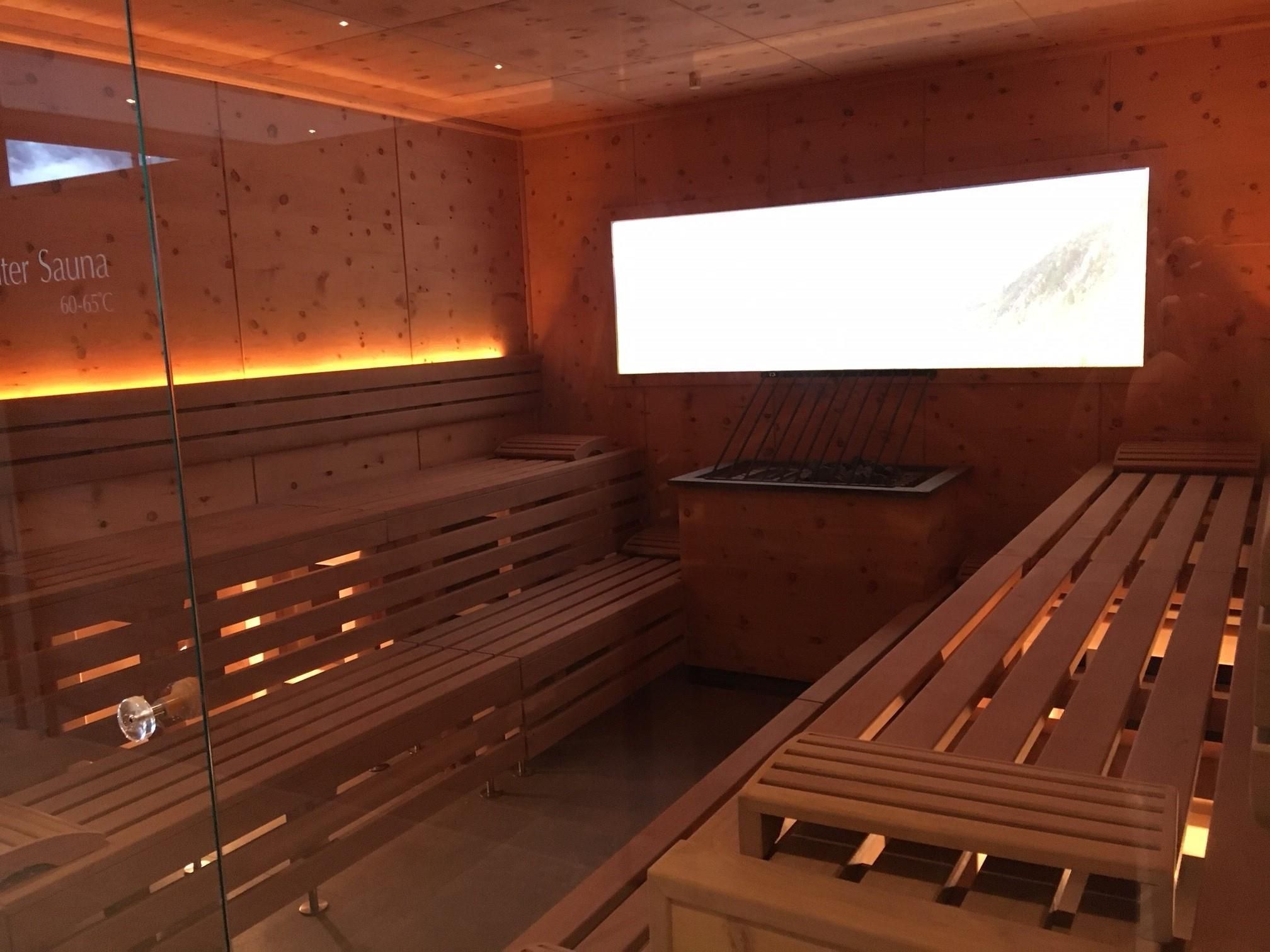 A modern sauna area with wooden loungers and soft lighting. In the background, a large screen is integrated, creating a relaxing atmosphere.