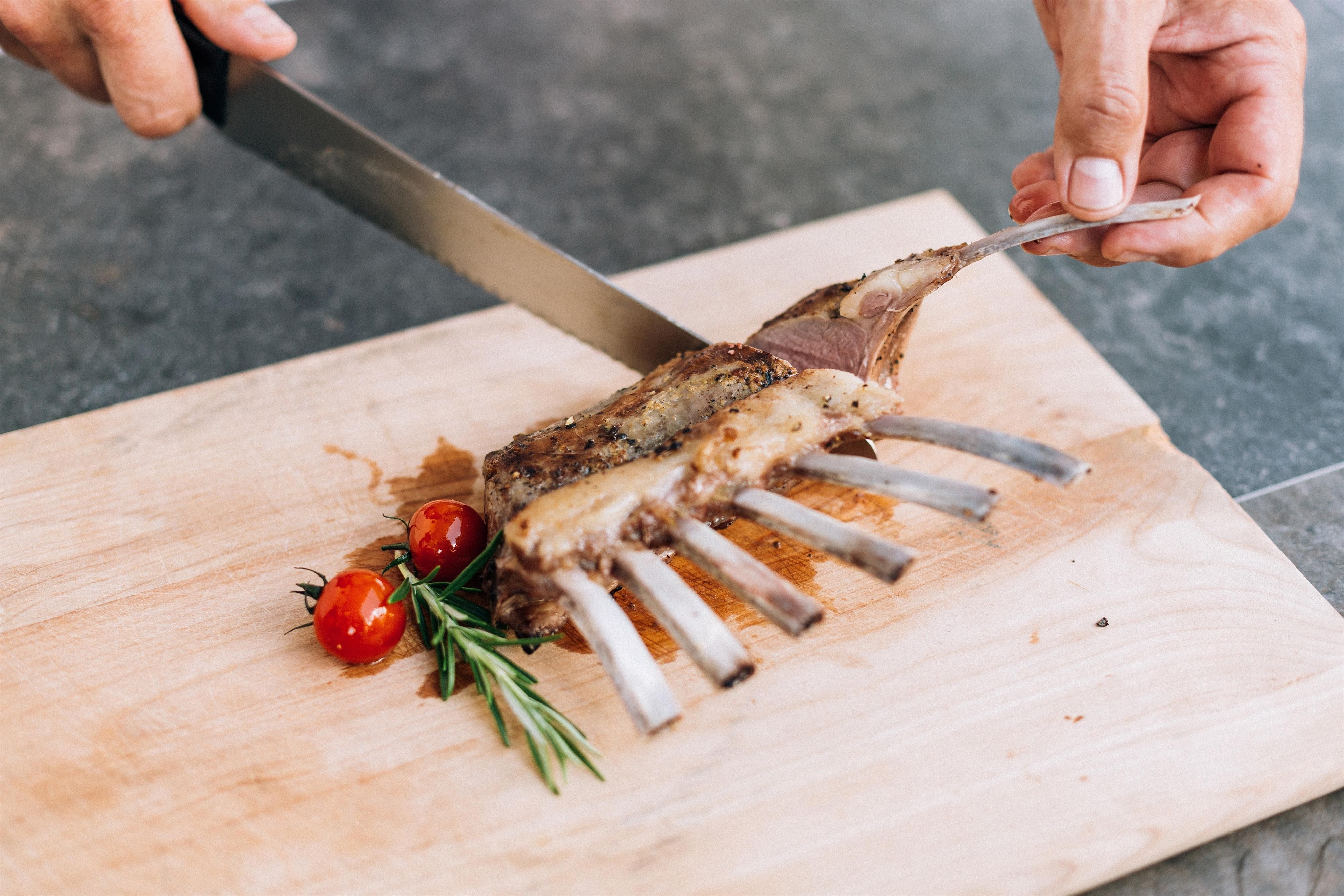 A chef is cutting a piece of meat on a wooden board. Next to it are cherry tomatoes and fresh rosemary.