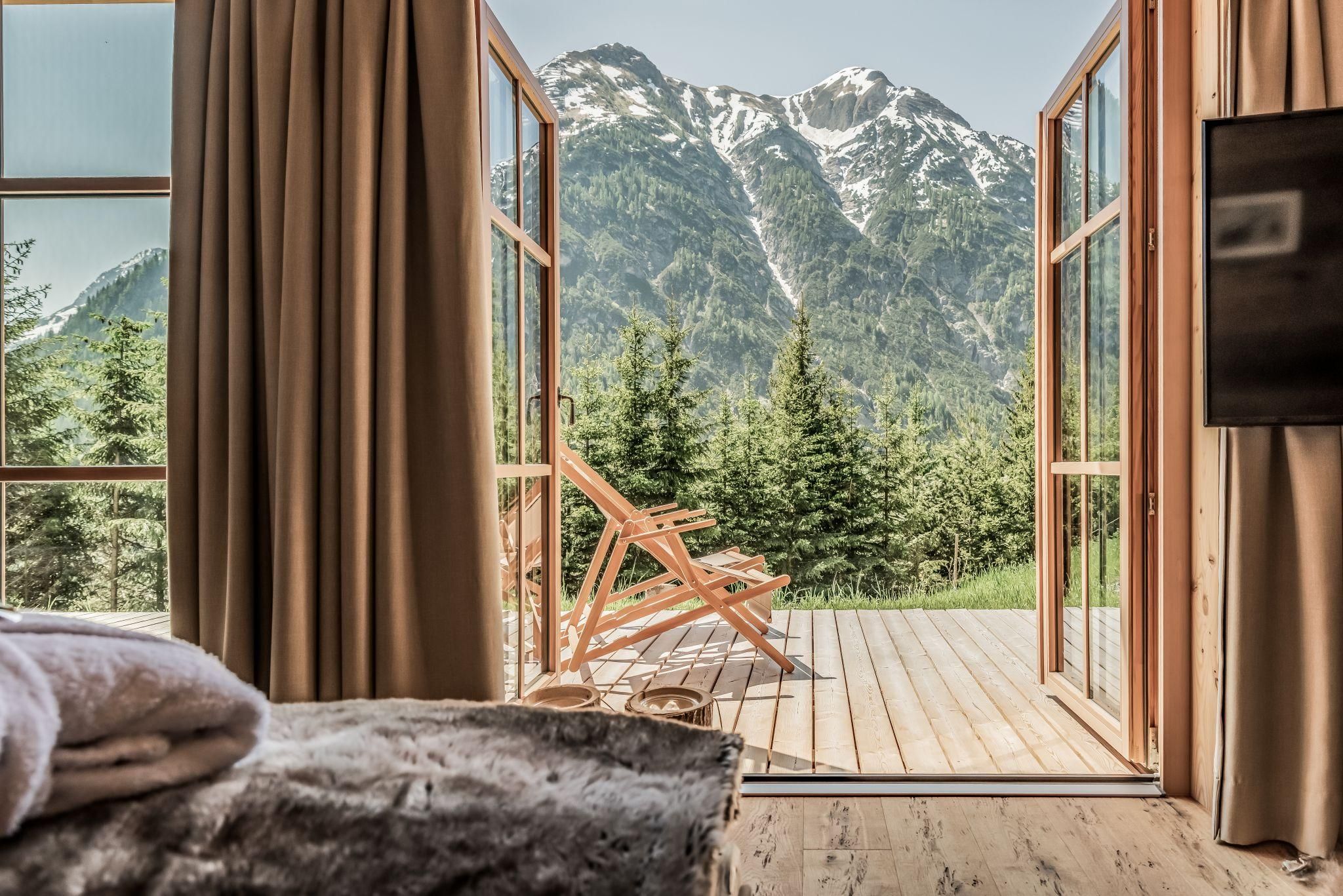 A cozy room with an open door leading to a terrace. In the background, majestic mountains and green trees can be seen.