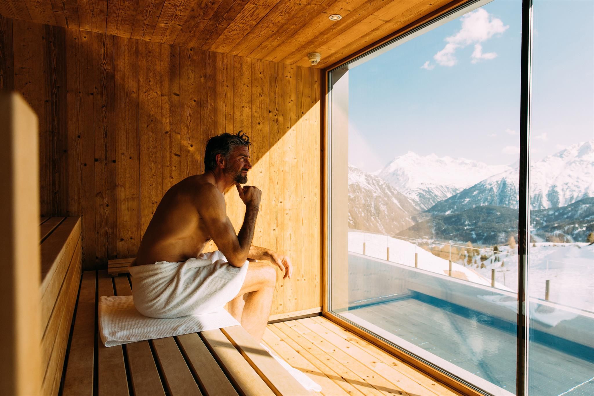 A man is sitting in a sauna and looking out the window at the snow-covered mountains. The atmosphere is relaxing and inviting.