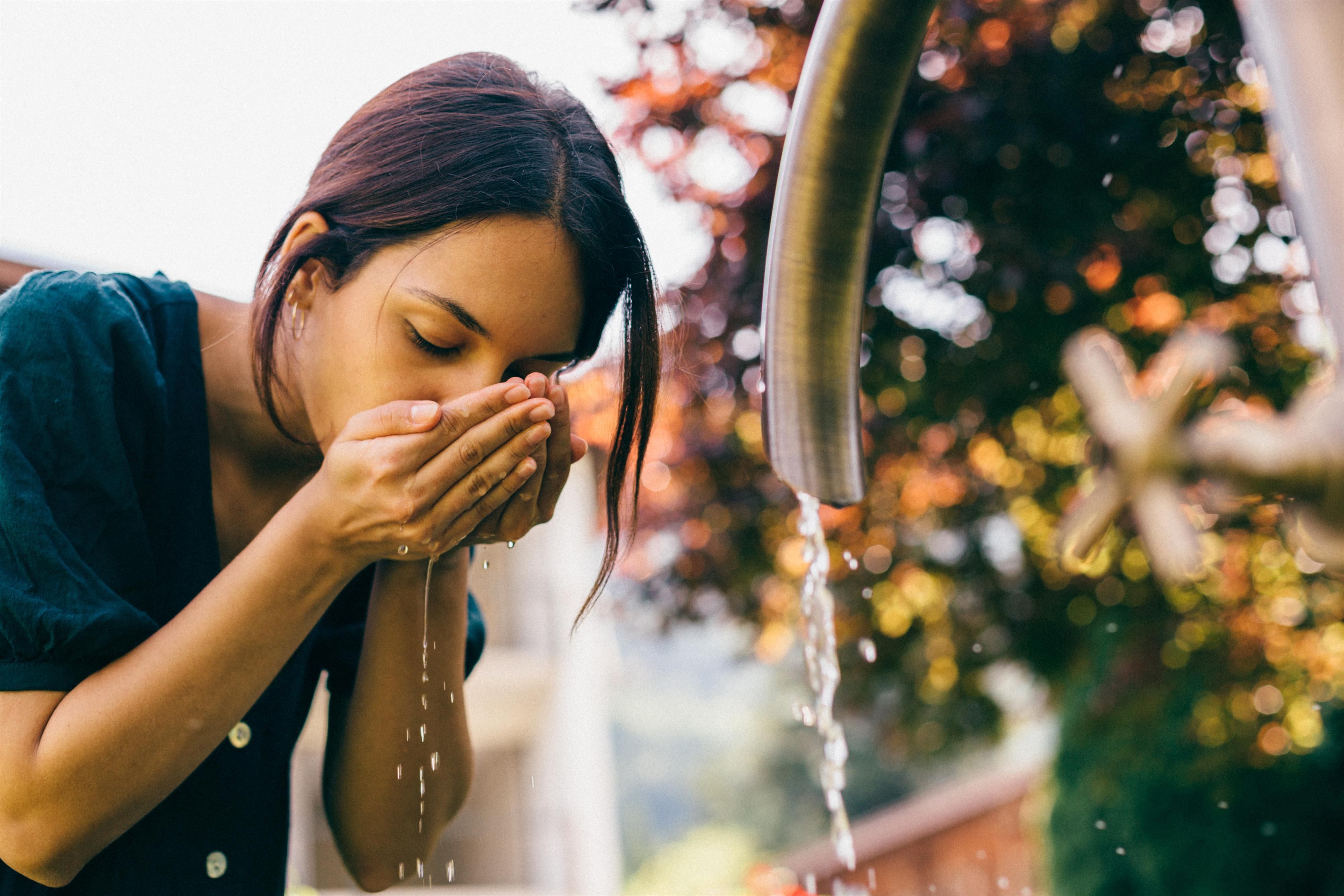 A woman leans over a faucet and washes her face. In the background, green trees and a blurred backdrop can be seen.