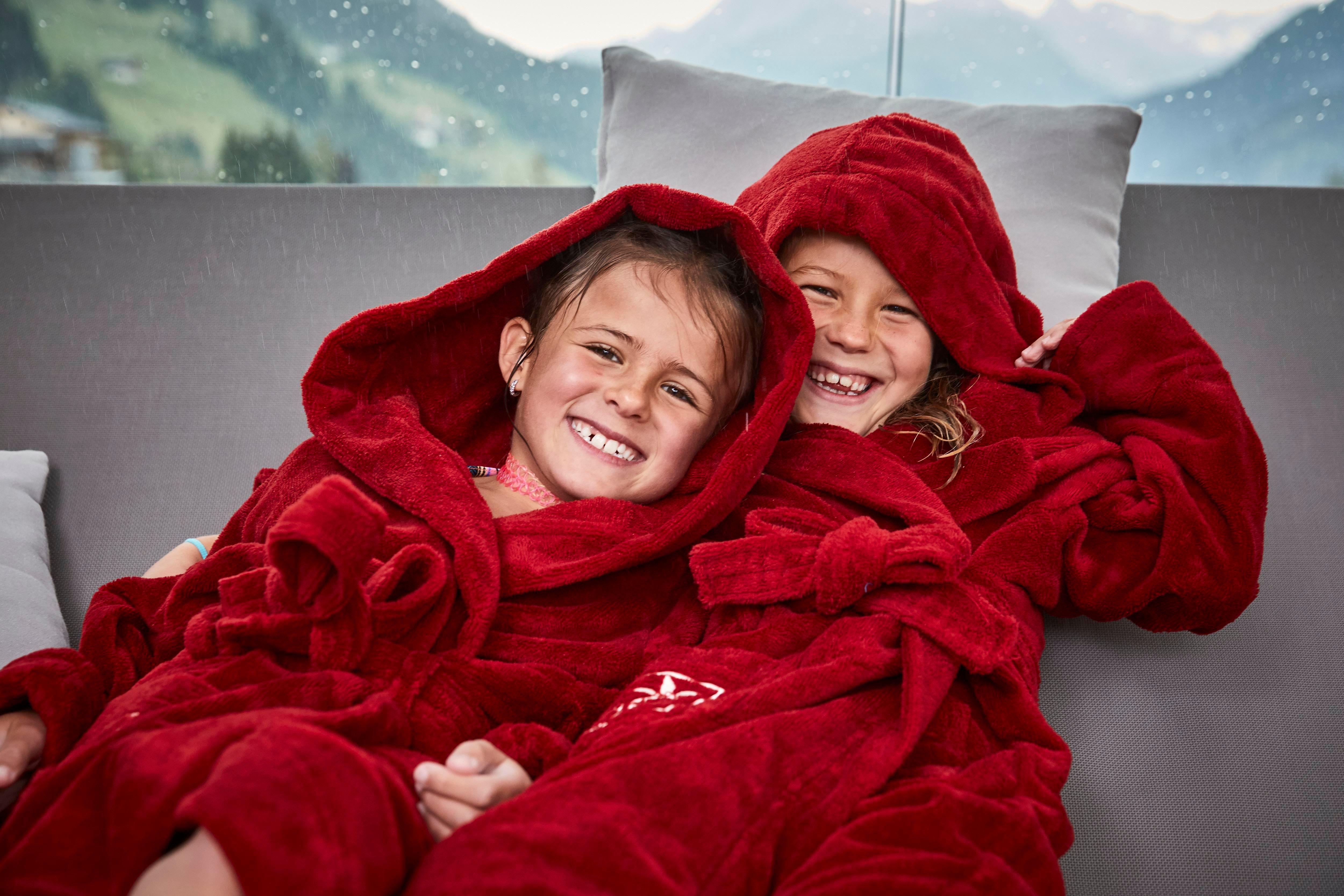 Two cheerful girls in red terry bathrobes sit next to each other. They smile relaxed and enjoy the view of the mountains.