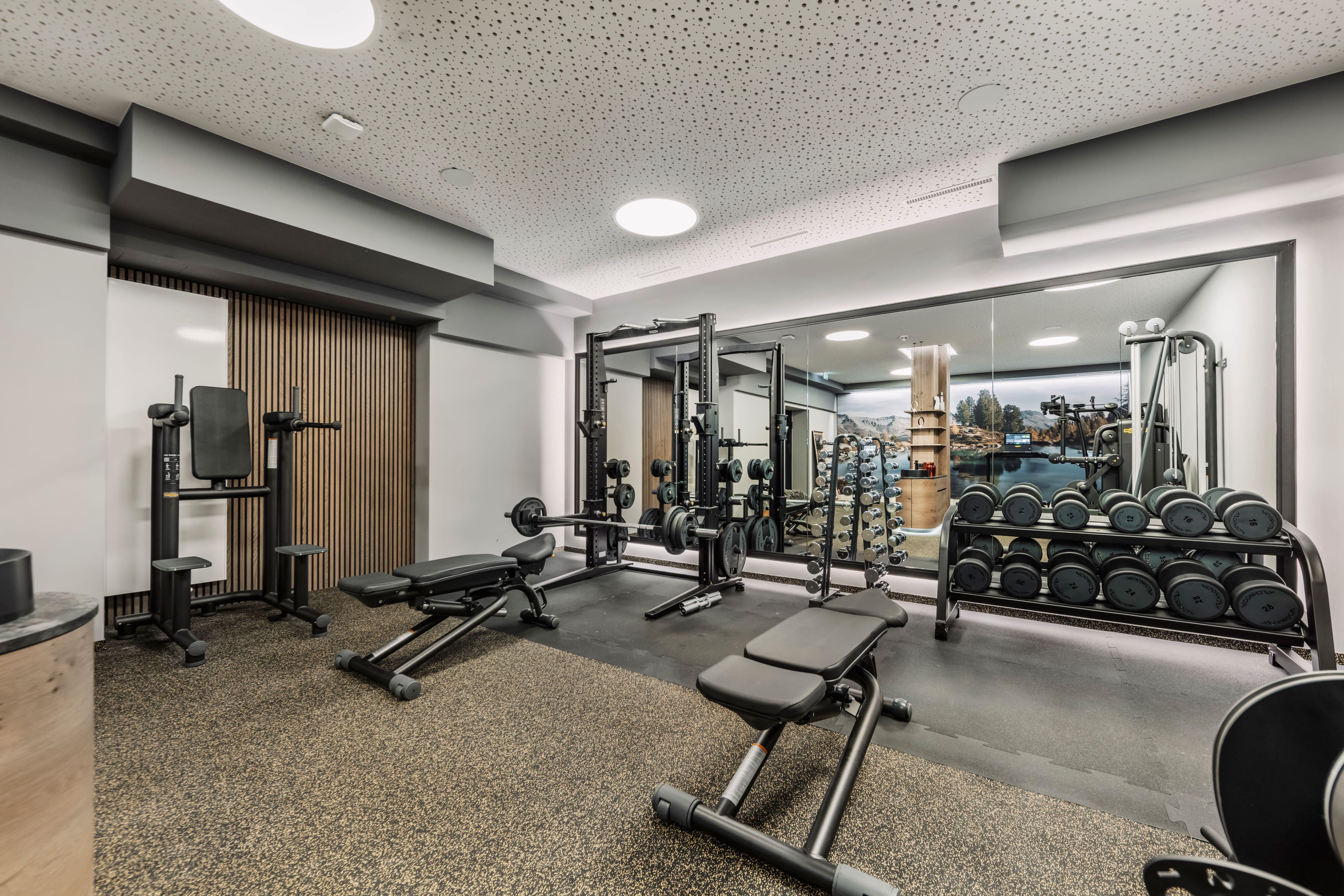 A modern gym with various training machines and weights. The walls are bright and mirrors reflect the room.