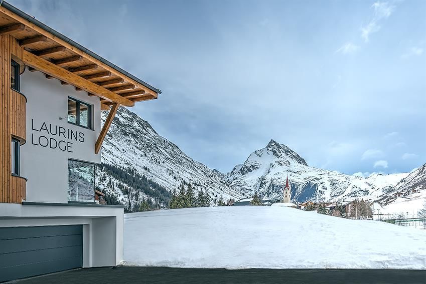 A modern lodge named "Laurins Lodge" in a snowy mountain landscape. In the background, majestic mountains can be seen under a cloudy sky.