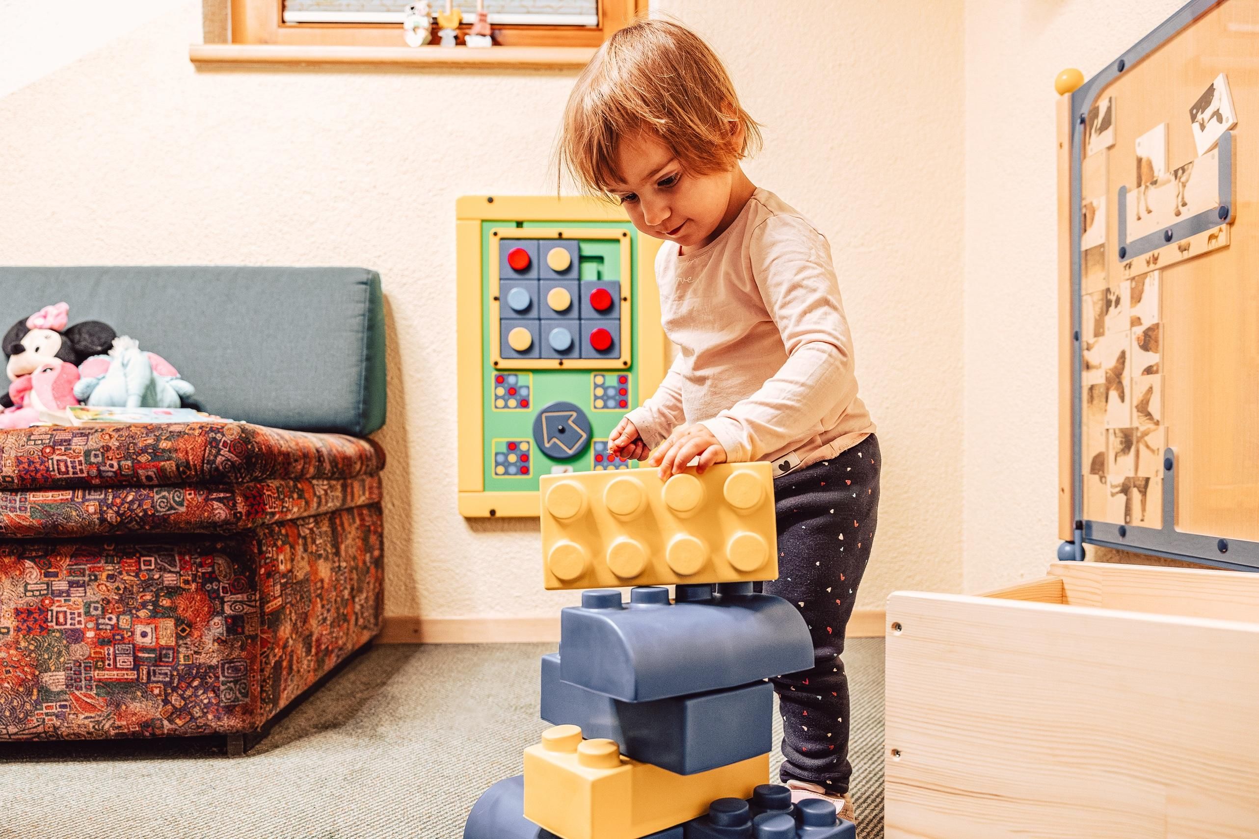 A small child is building with large, colorful building blocks. In the background, a sofa and an interactive game board are visible.