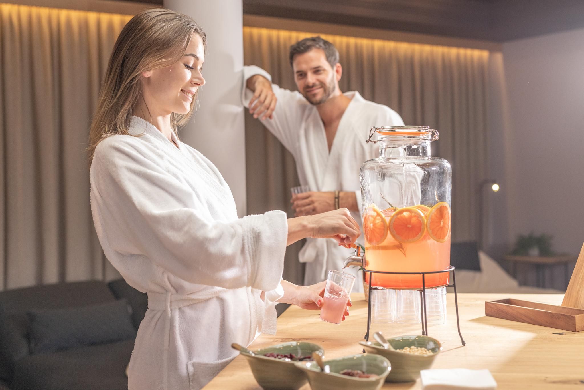 A woman in a bathrobe is pouring a refreshing drink from a dispenser, while a man in the background is smiling. On the table, snacks and fresh fruit can be seen.
