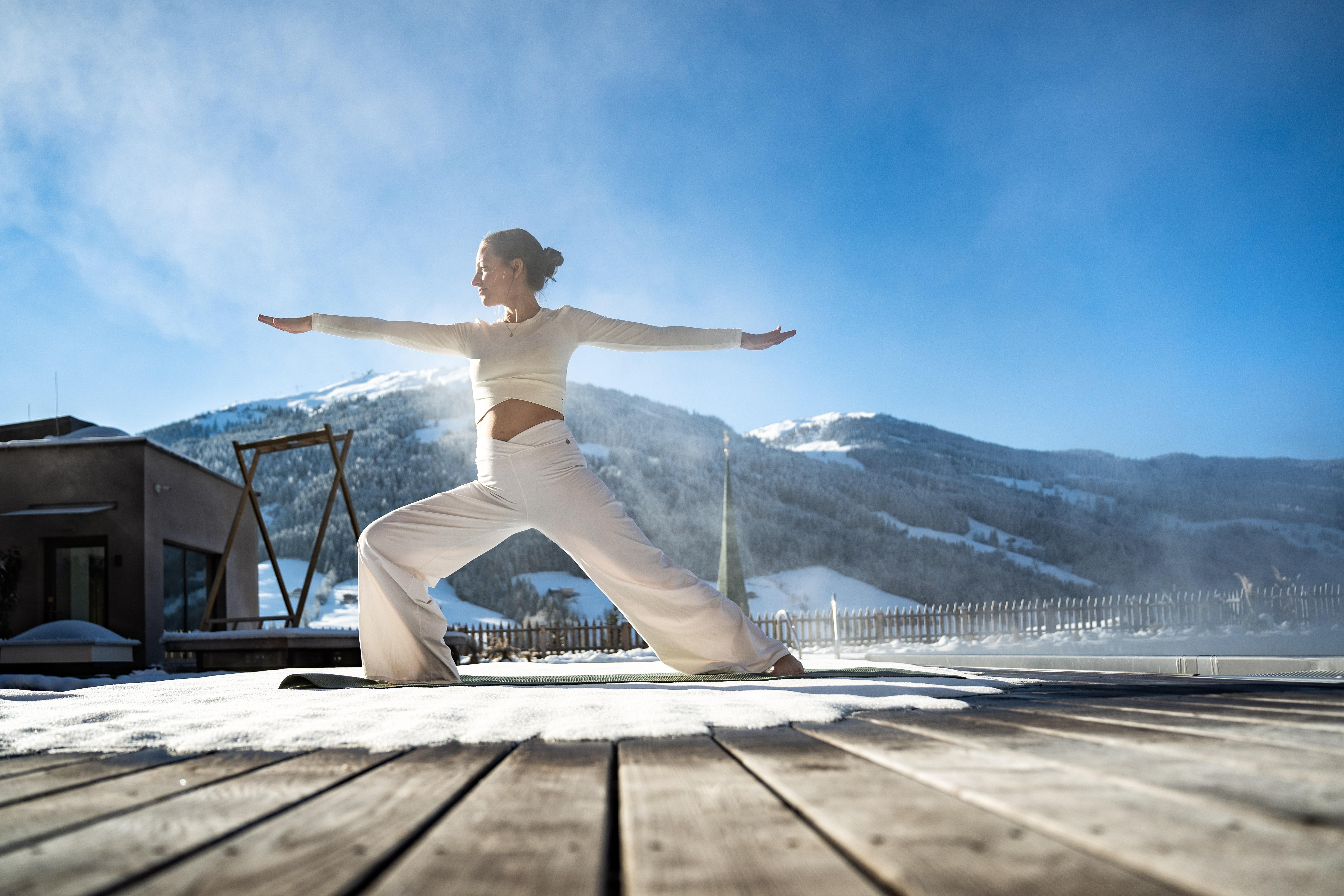 A woman practices yoga on a wooden terrace in the snow. In the background, there are snow-covered mountains and a clear blue sky.