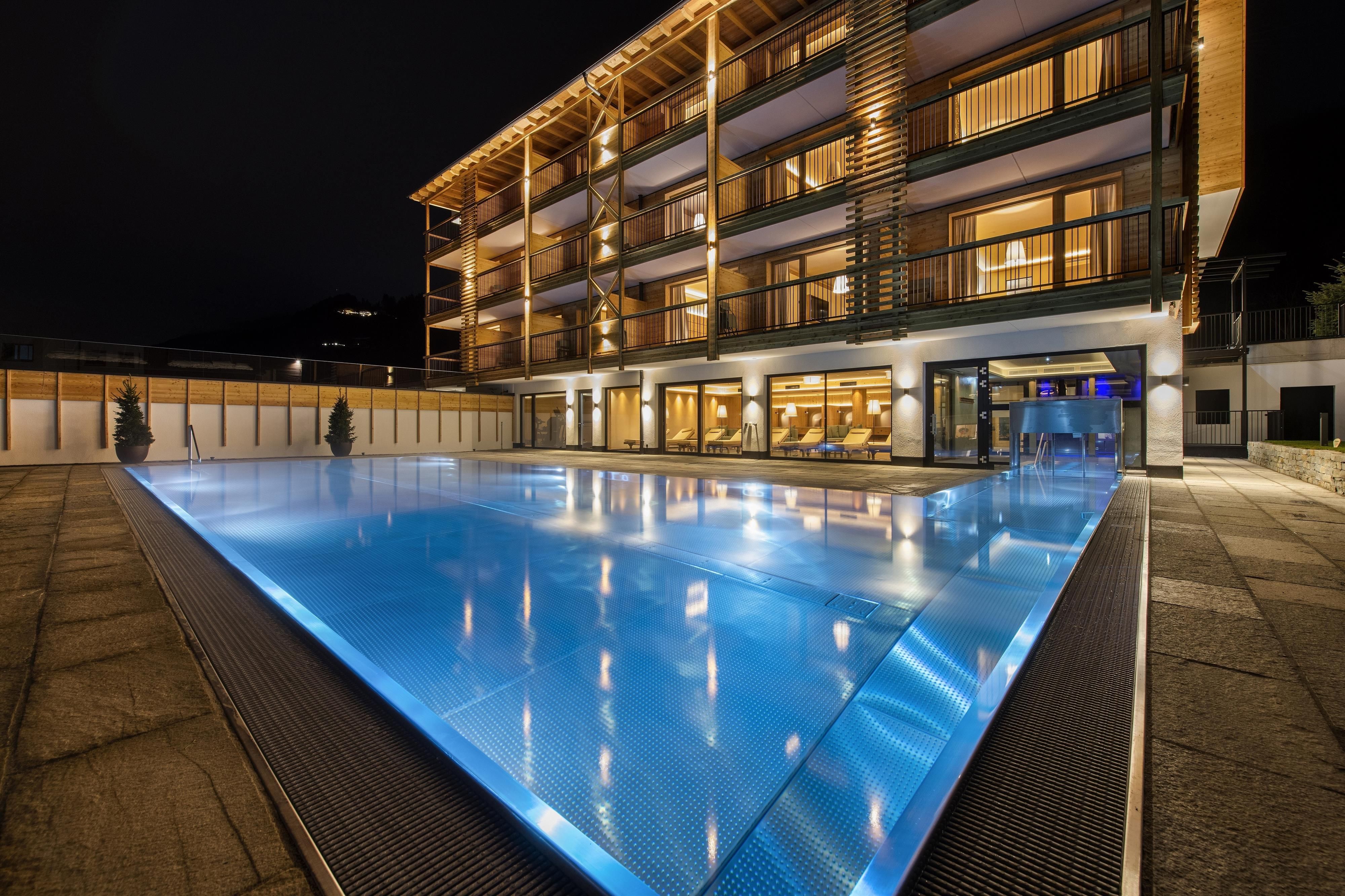 A modern hotel building with an elegant outdoor pool. The pool radiates in a clear, blue light at night.
