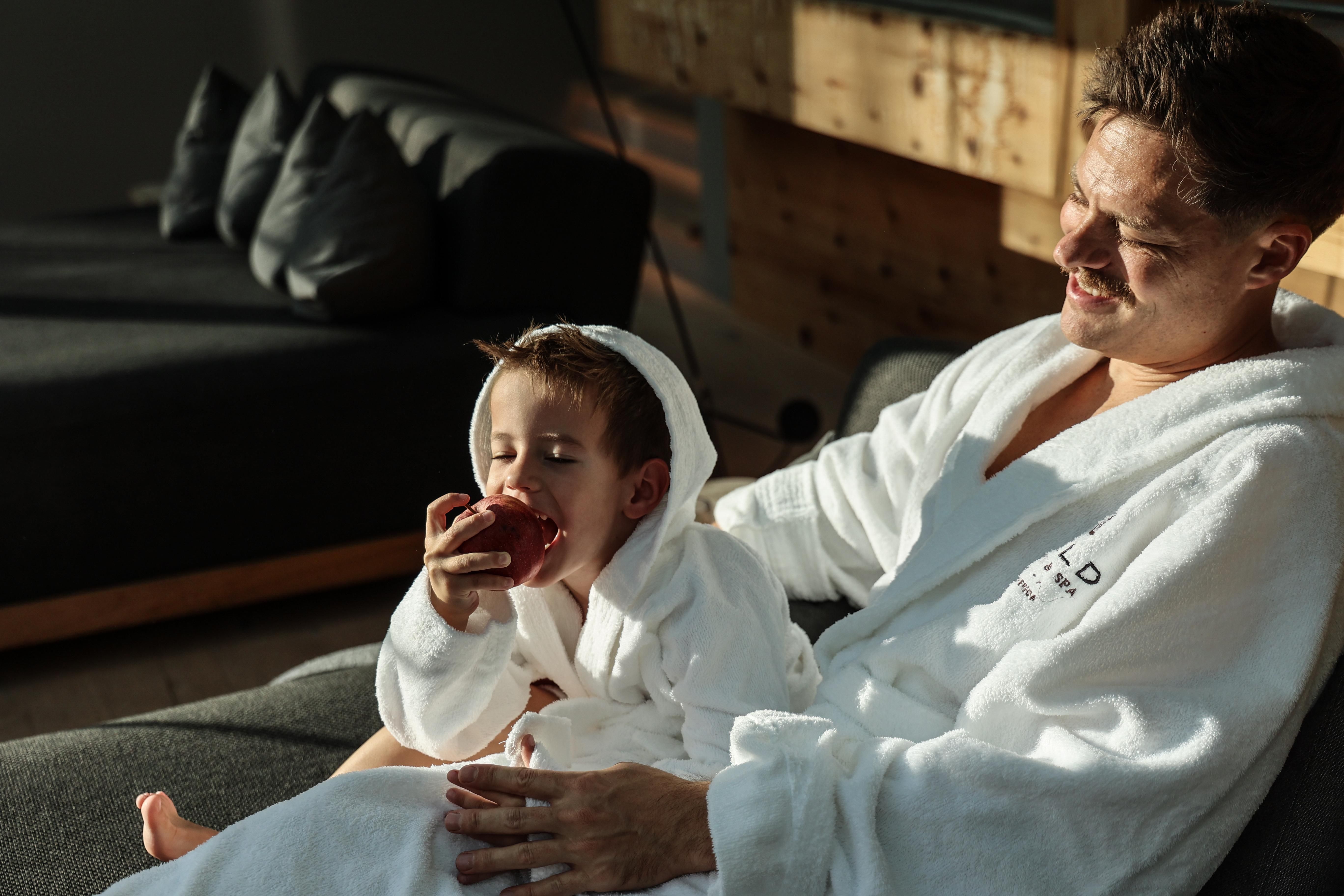 A father and his son are comfortably sitting on a couch in bathrobes. The boy is biting into an apple and both are smiling relaxed.