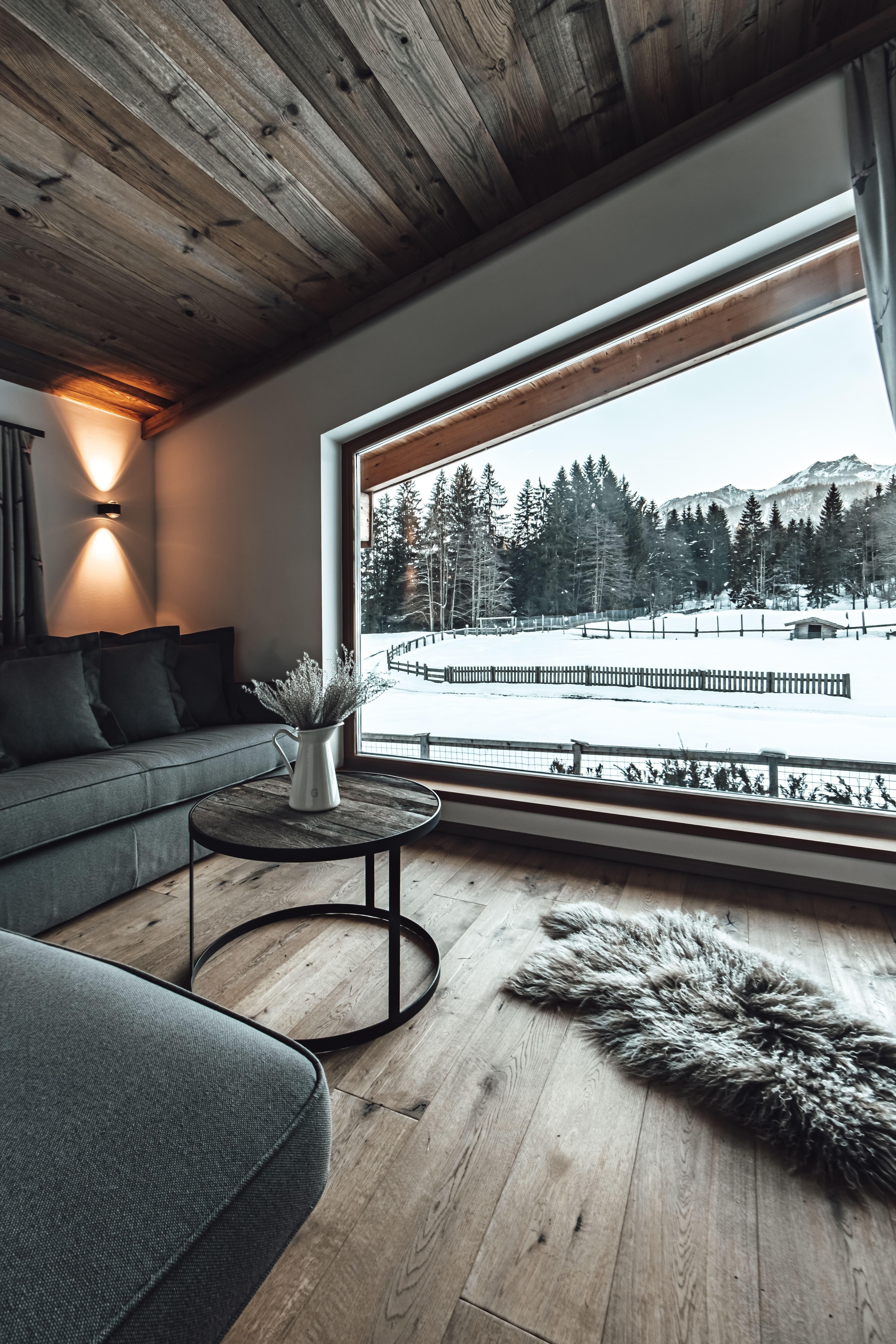 A cozy living room with a dark sofa and a round table. Through the large window, there is a view of snow-covered trees and mountains.