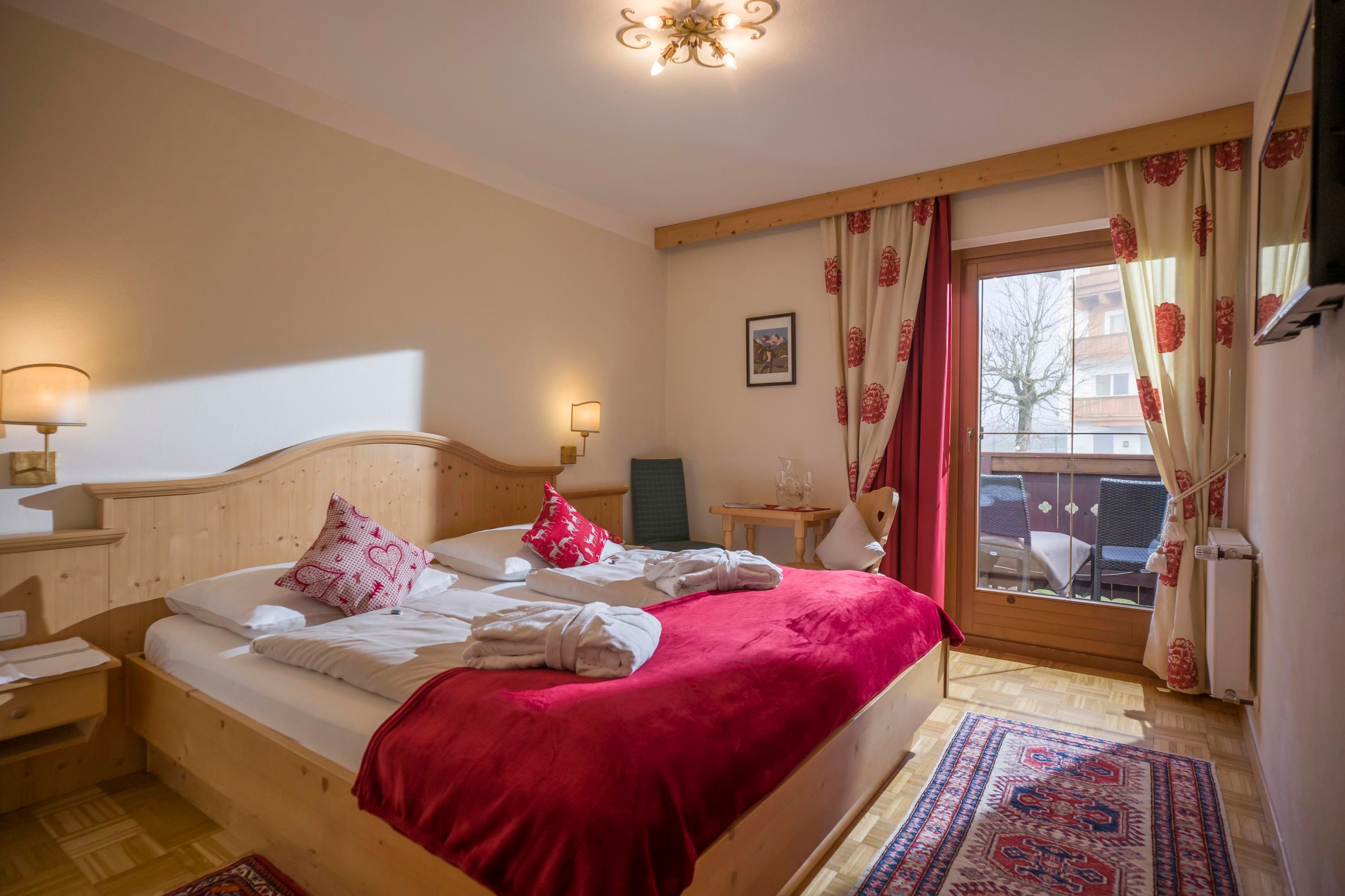 A cozy bedroom with a double bed and red pillows. There is a terrace with a view and bright curtains.