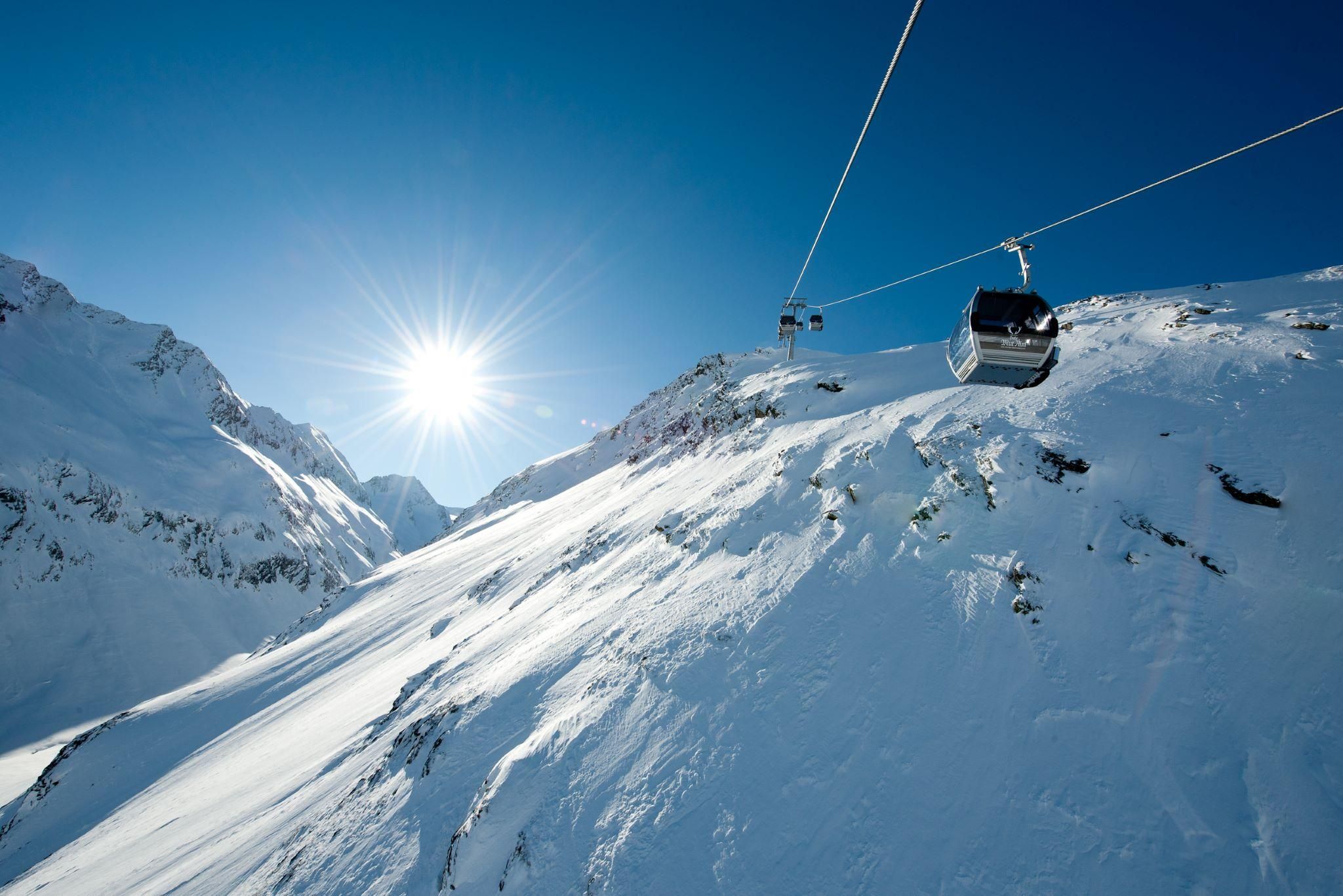 A cable car travels over snow-covered mountains under a bright blue sky. The sun shines brightly, highlighting the winter landscape.
