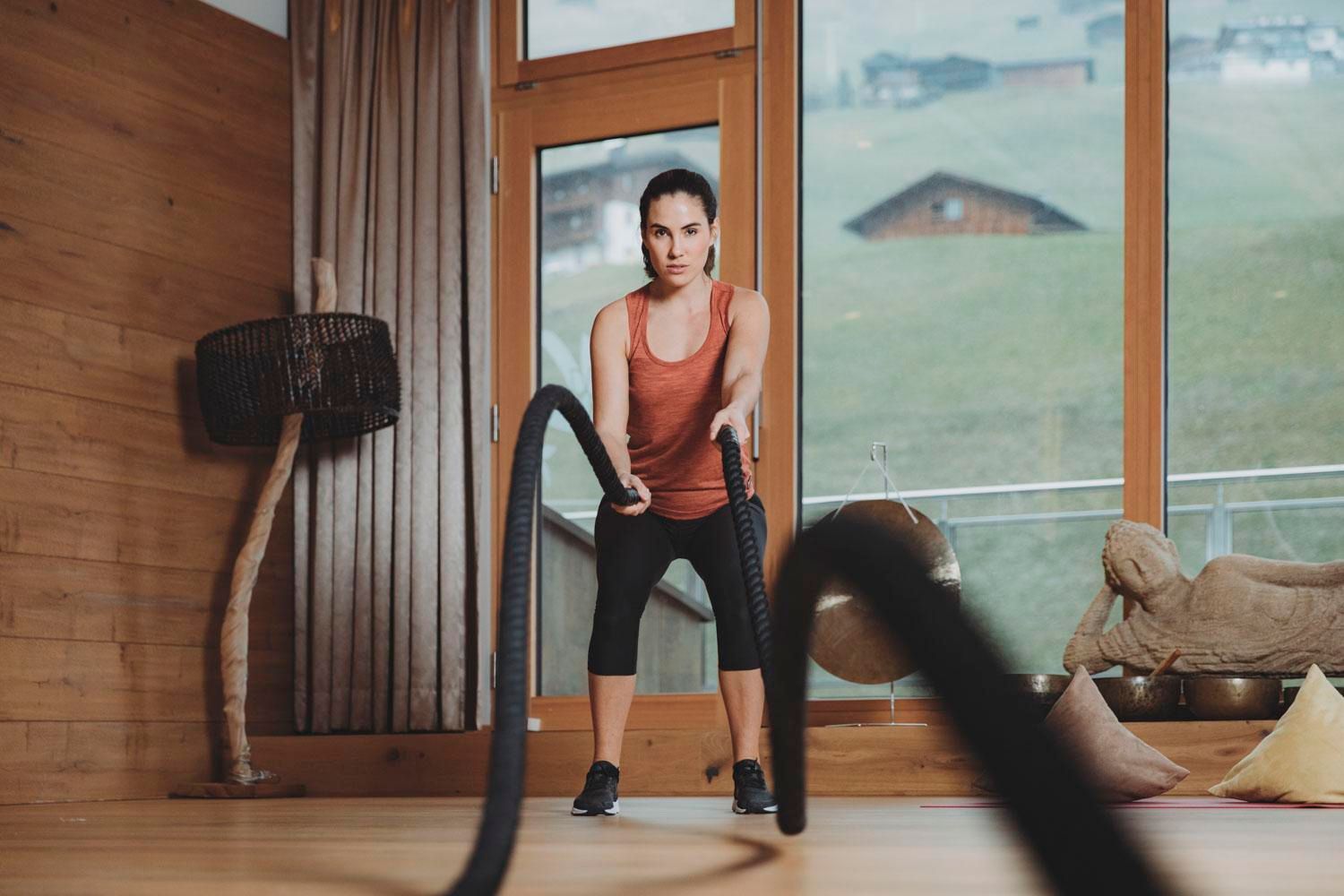 A woman is doing fitness with ropes in a modern room. A green landscape is visible through the windows.