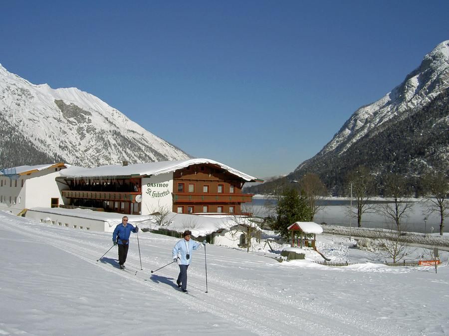 An idyllic winter landscape with two people cross-country skiing. In the background, snow-covered mountains and a building can be seen.