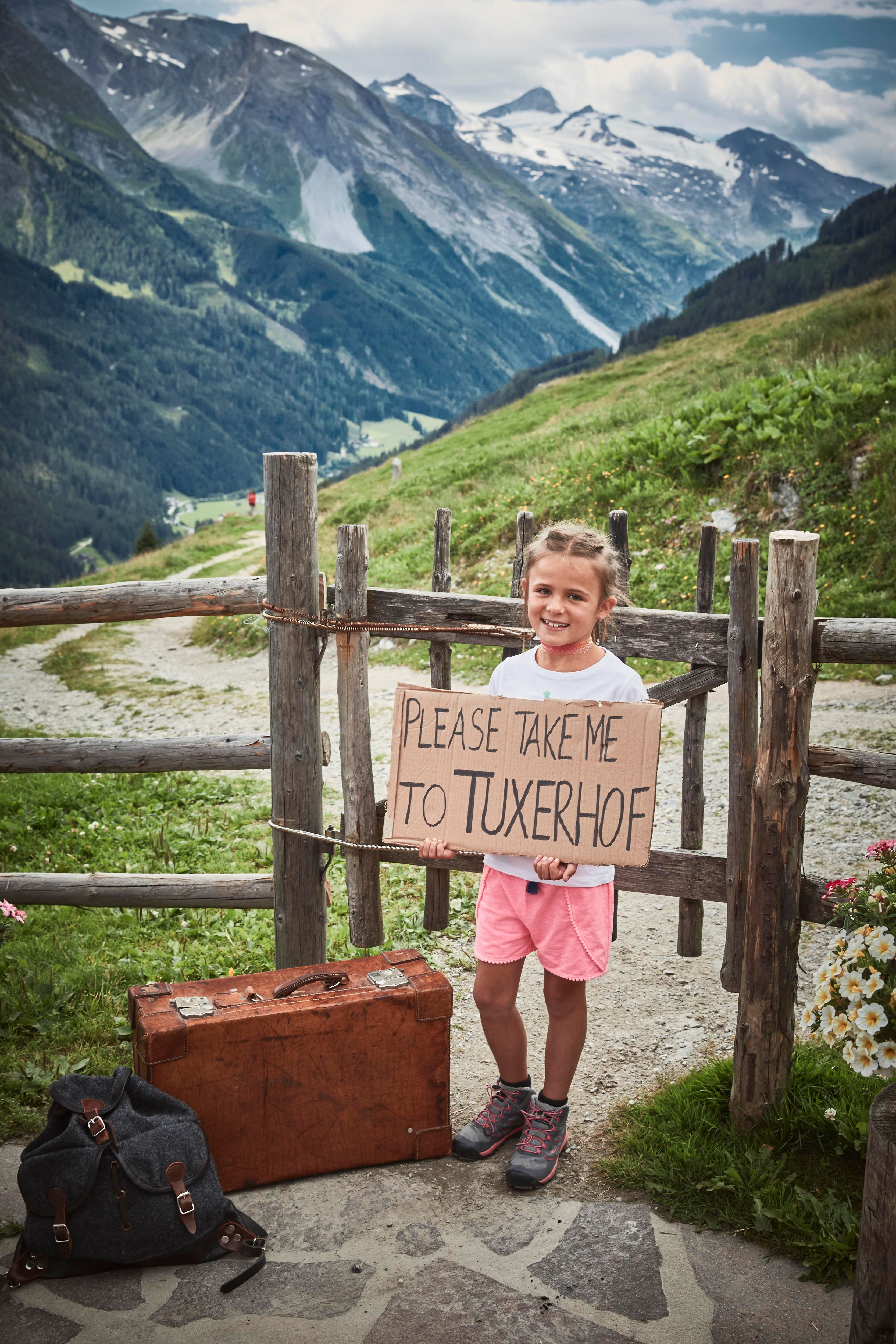 A girl is holding a sign that says "Please take me to Tuxerhof". In the background, mountains and a green landscape can be seen.