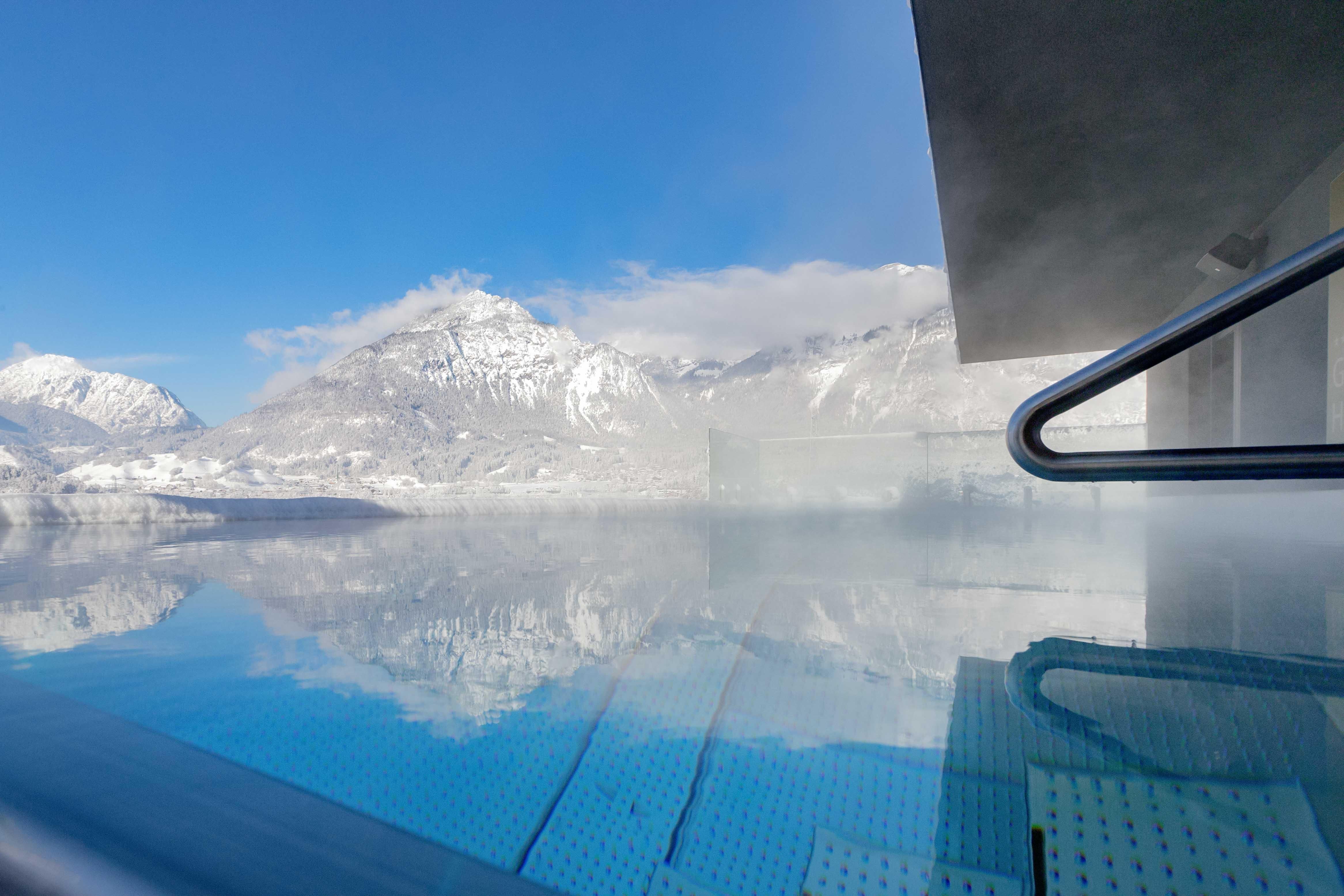 An infinity pool with a view of snow-capped mountains beneath a clear blue sky. The calm water surface reflects the surrounding mountain landscape.