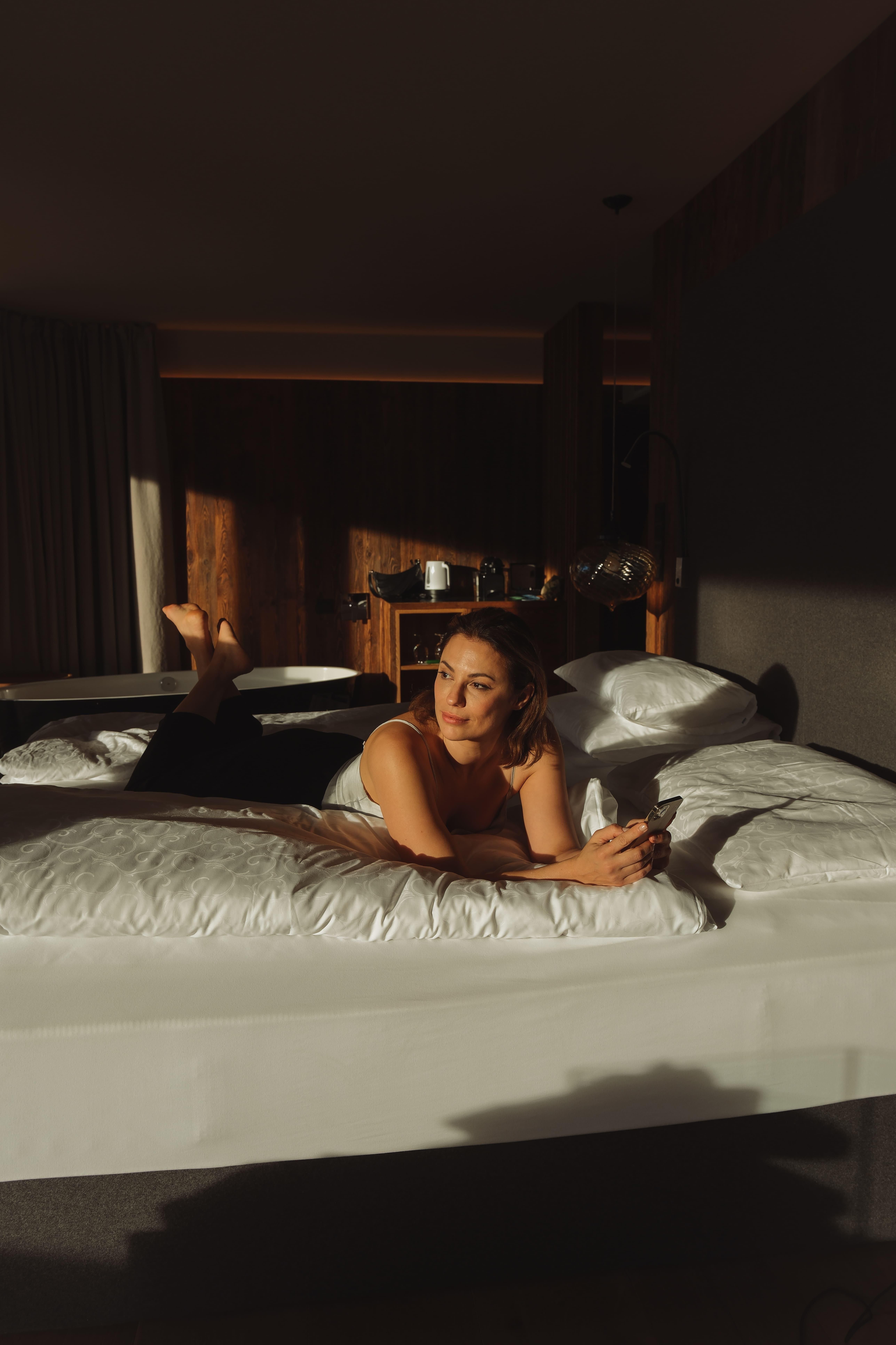 A young woman relaxes on a modern bed and looks at her smartphone. The room has warm light and a cozy atmosphere.