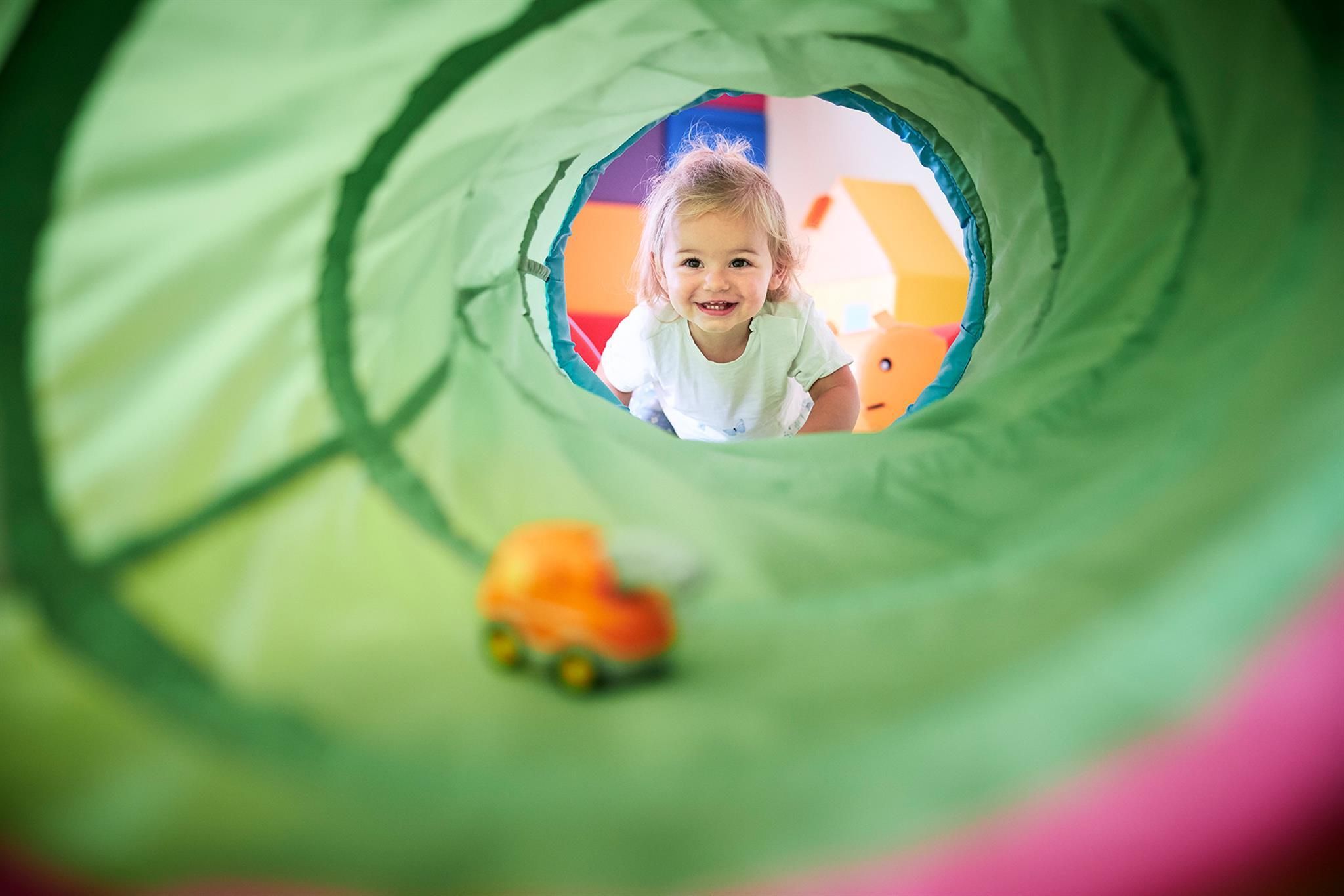A small child is happily smiling through a green play tunnel. In front of them lies a colorful toy car.