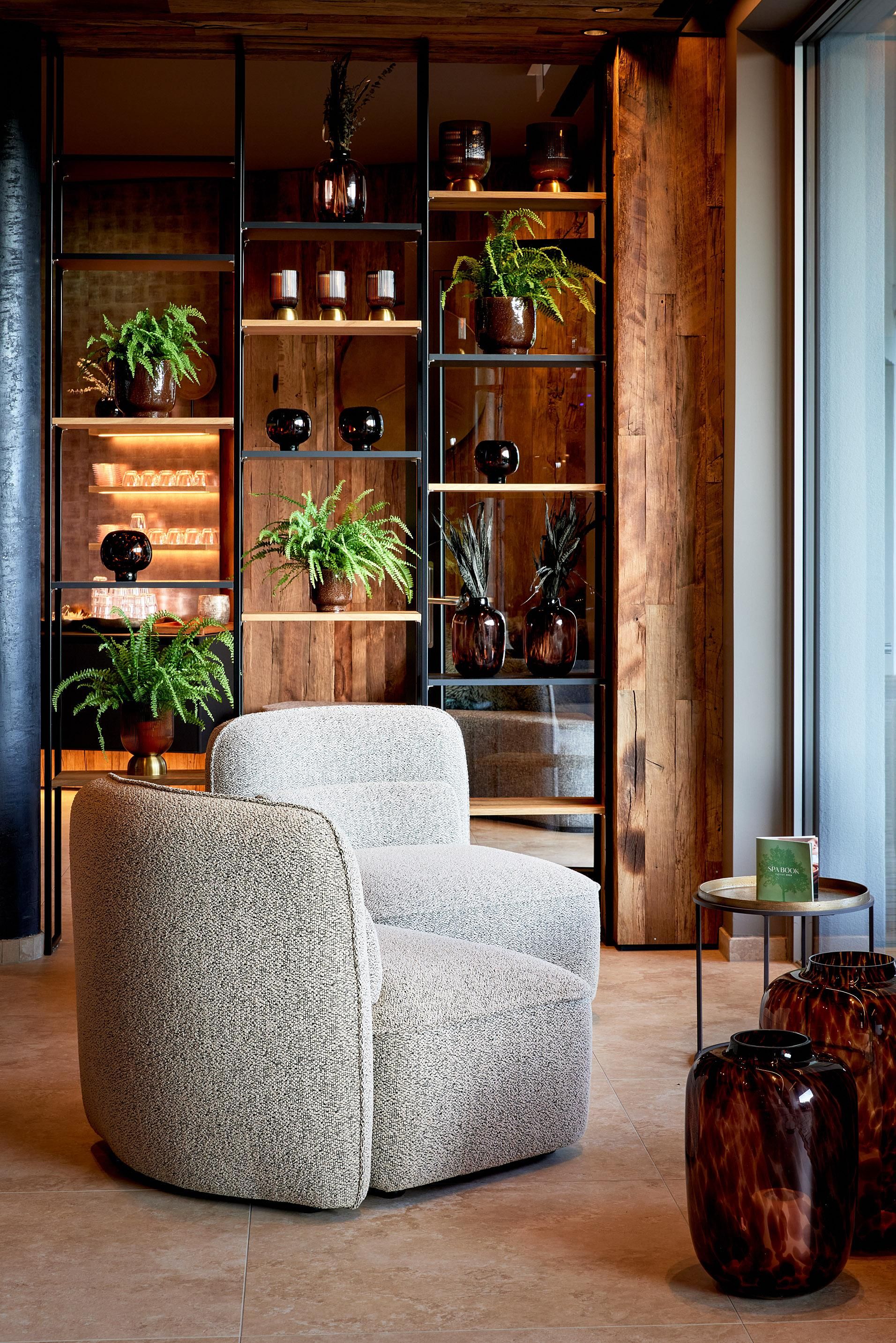 A modern armchair stands in front of an elegant shelf with plants and decorative objects. The warm wood tones and stylish design create a cozy atmosphere.