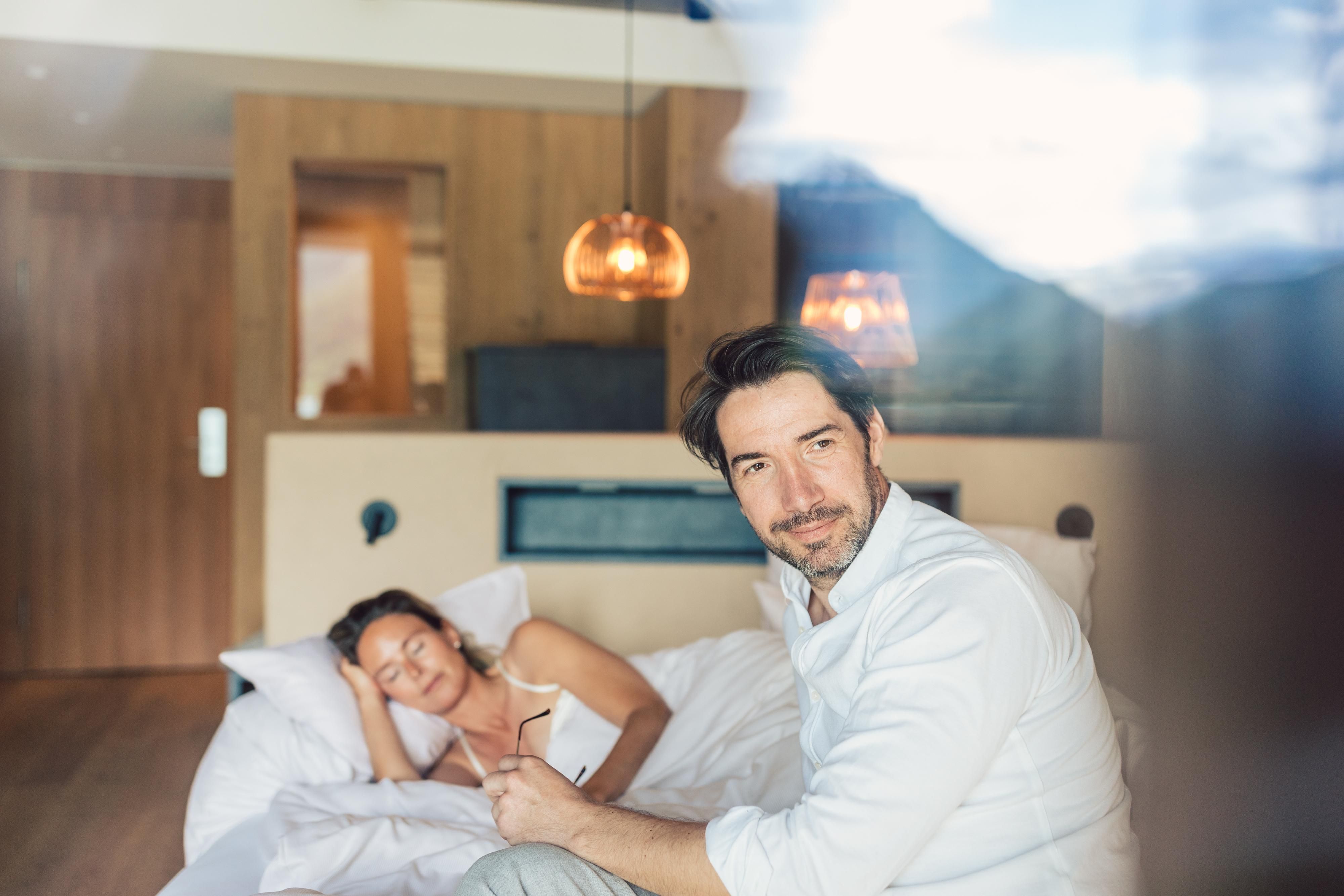 A man sits smiling on the bed while a woman sleeps beside him. The room has a modern, cozy atmosphere with wooden details and warm light.