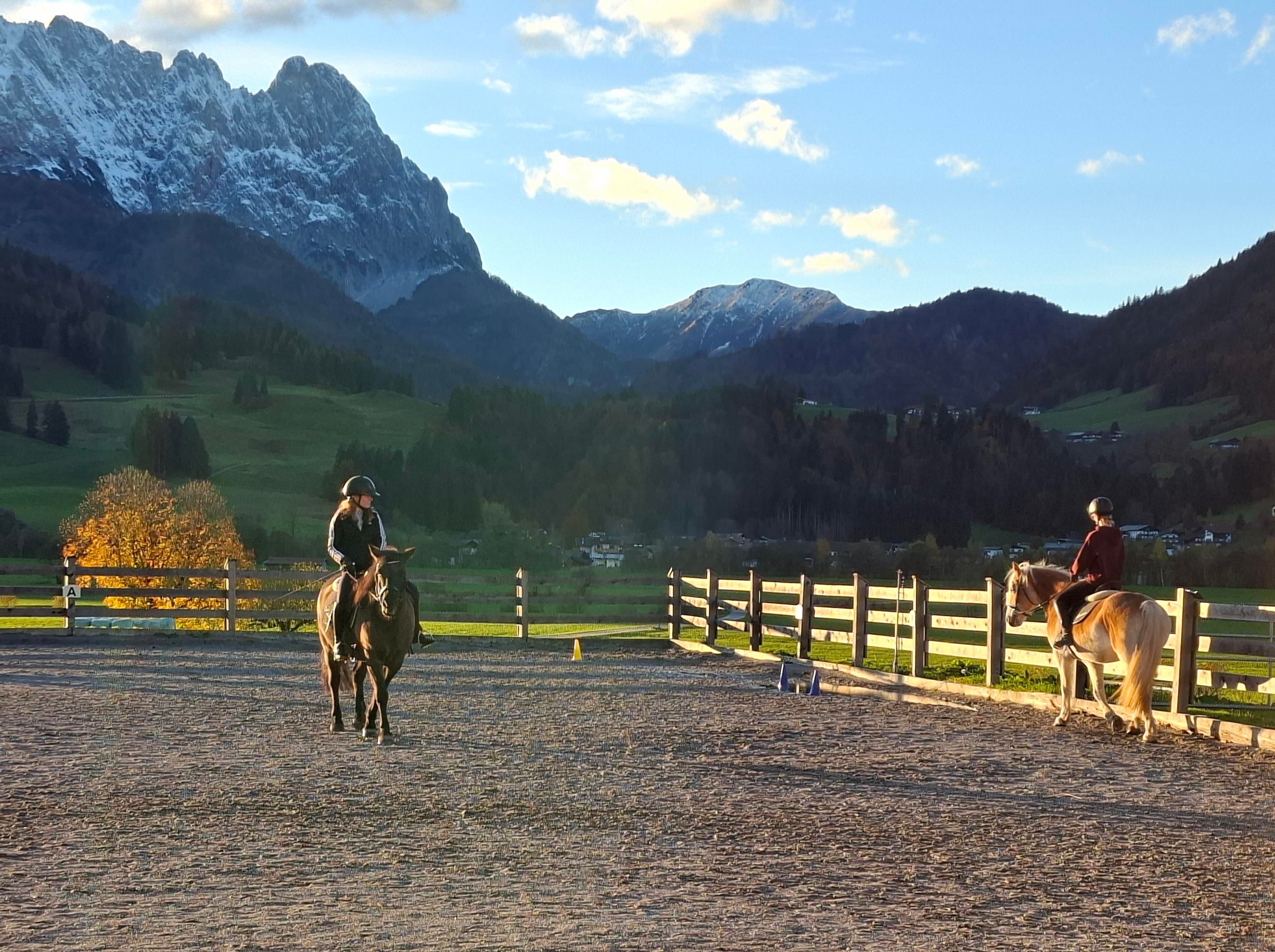 Two horse riders in a riding arena with beautiful mountain scenery in the background. The sun is shining and the surroundings are calm and picturesque.