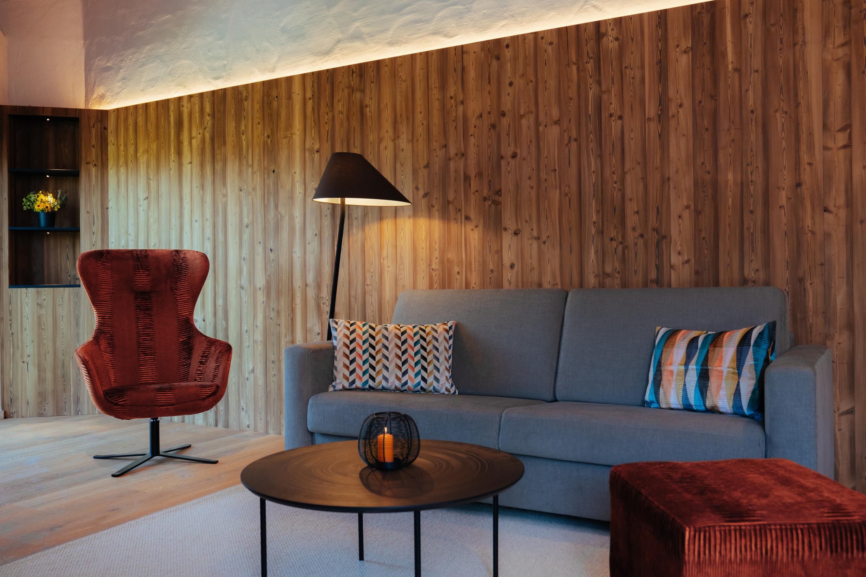 A modern living room with a gray sofa and a red armchair. Warm wooden walls and soft lighting create a cozy atmosphere.
