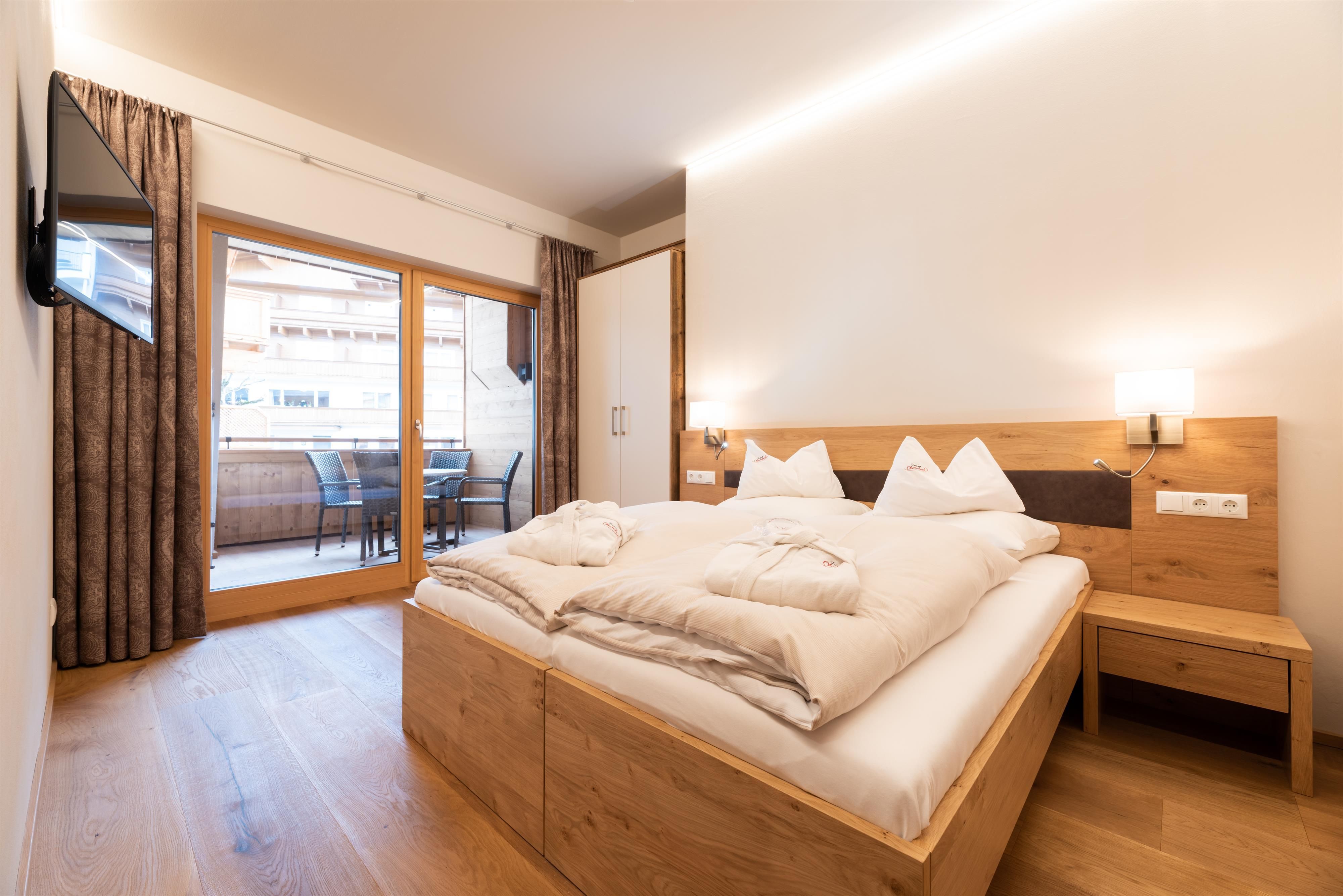 A modern bedroom with a large, comfortable bed and wood paneling. The room has access to a balcony with seating and plenty of natural light.