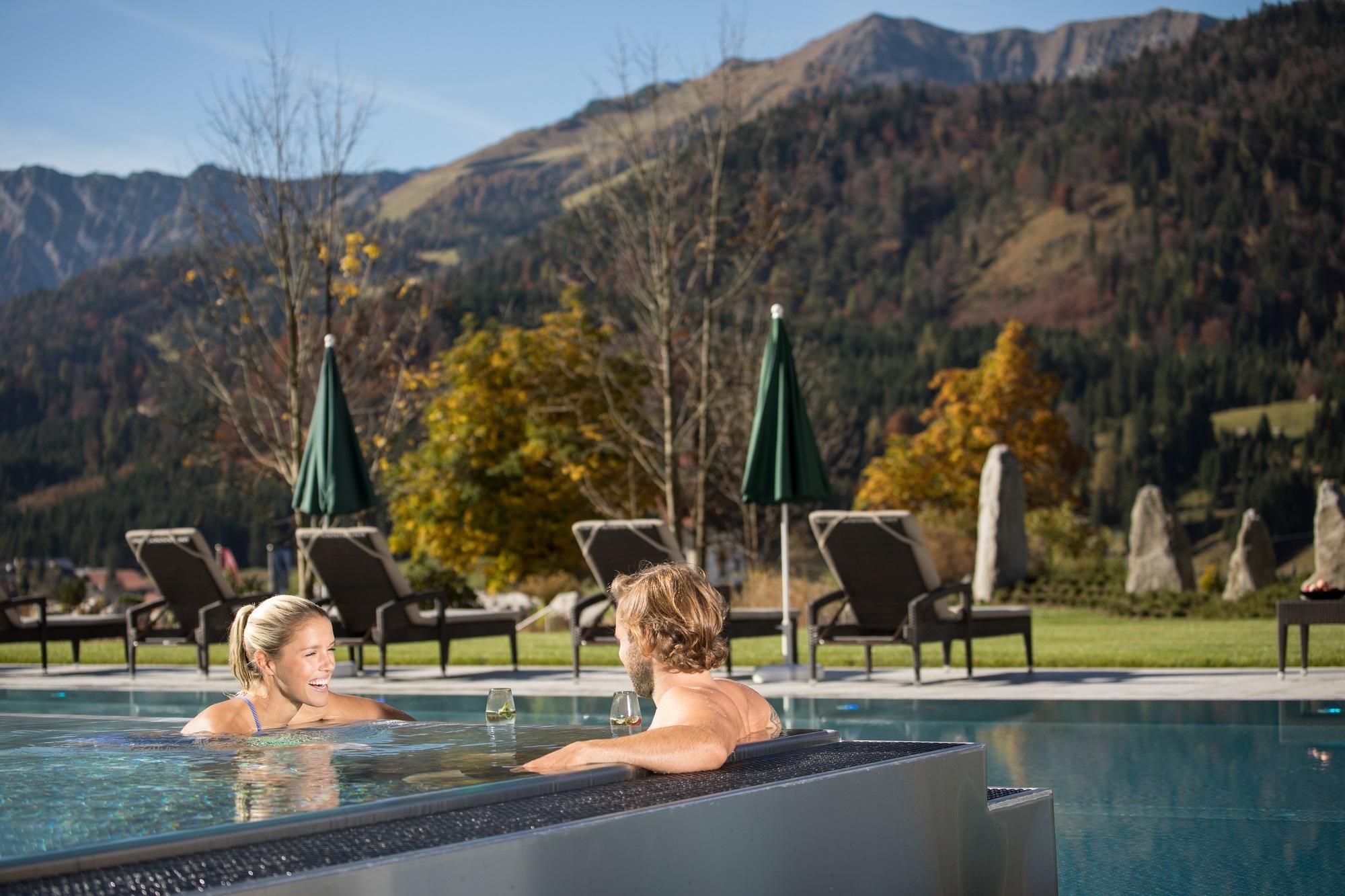 A couple relaxes in the warm water of an outdoor pool with a view of the mountains. In the background, there are lounge chairs and shade-providing sun umbrellas.