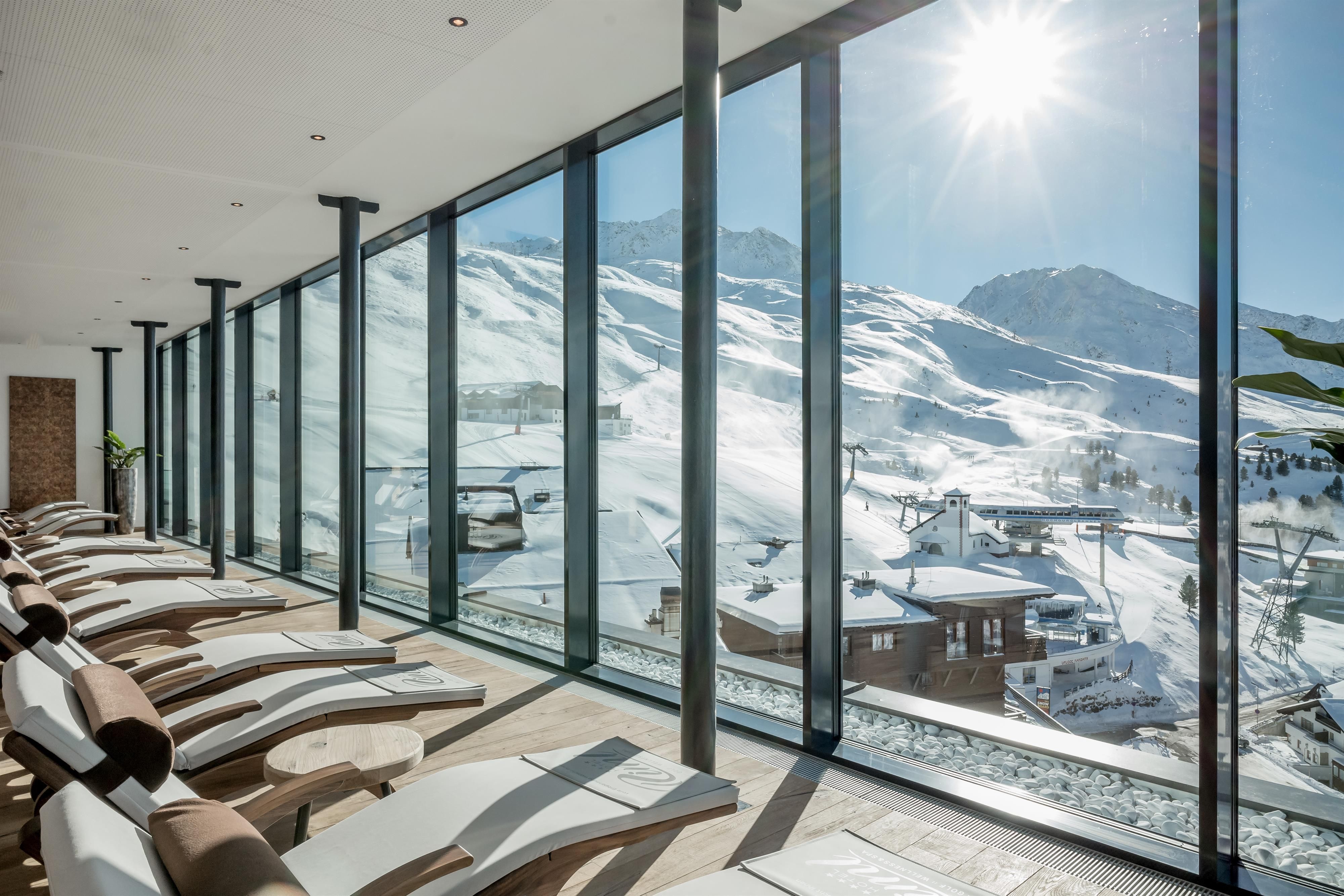 A bright wellness area with loungers and large windows. Outside, you can see snow-covered mountains and a sunny landscape.