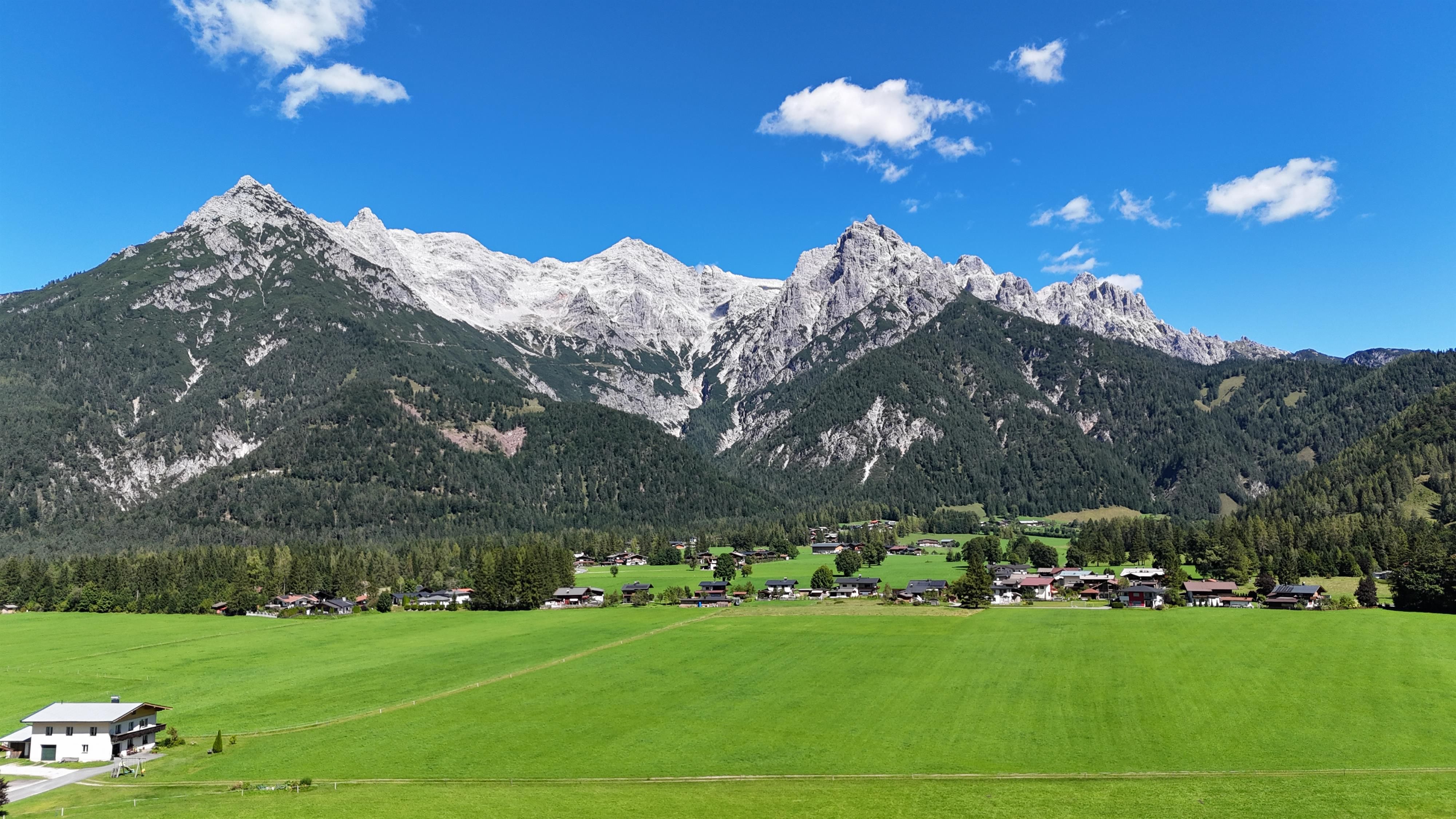 An impressive mountain landscape with high peaks and green valley floor. In the foreground, meadows and small houses are visible.