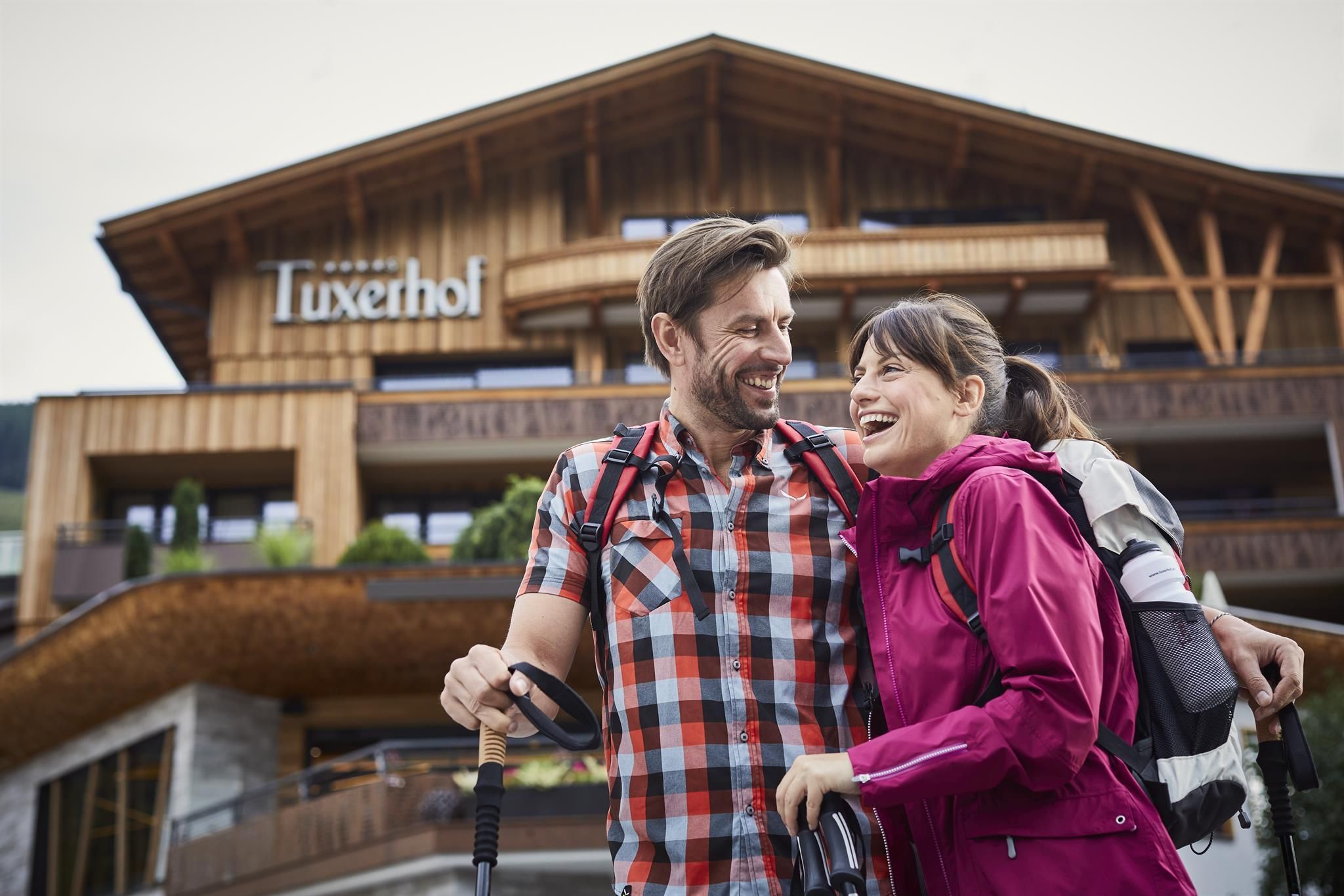 A happy couple stands in front of a hotel with wooden elements. Both are wearing hiking gear and are smiling friendly.