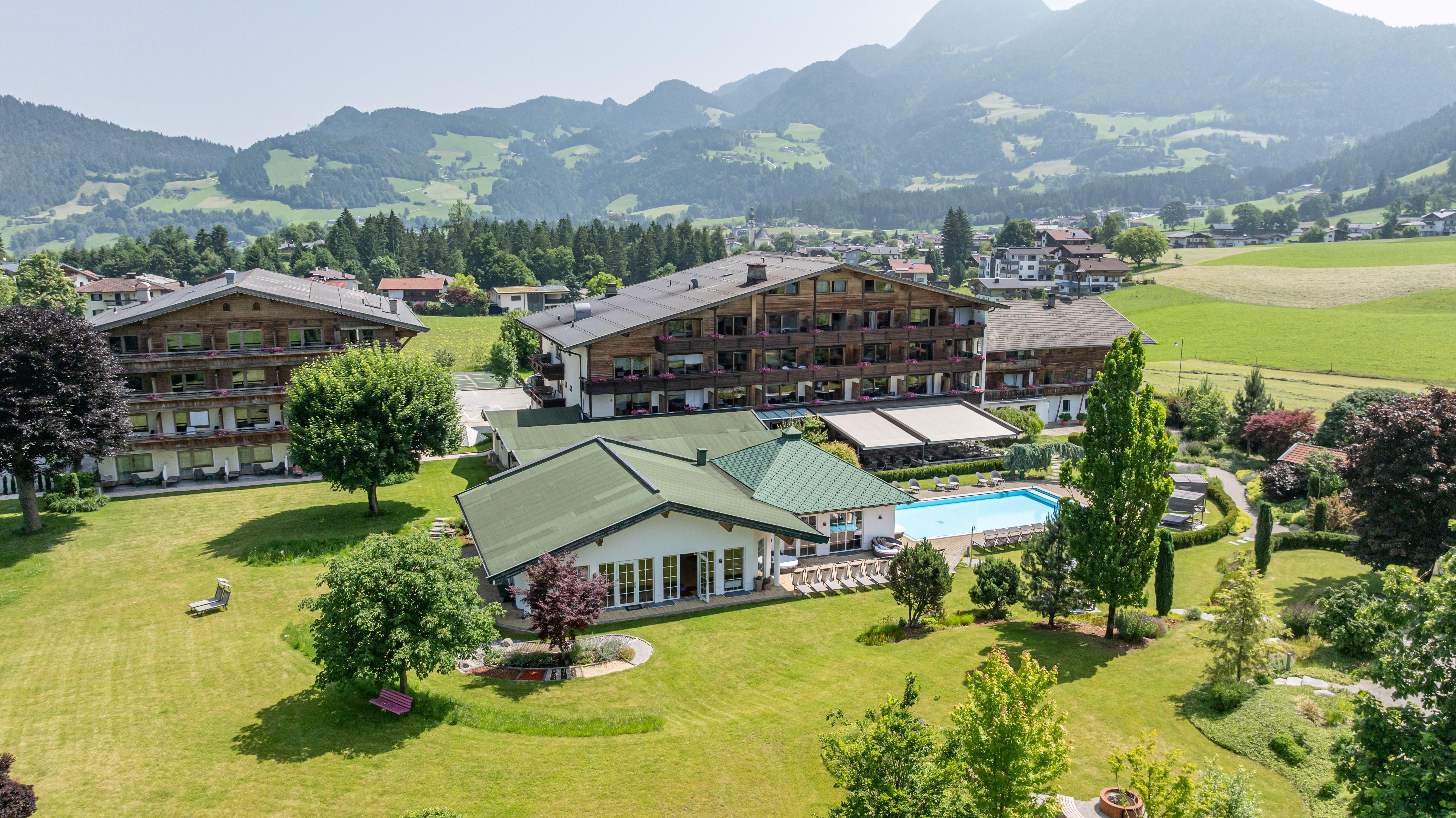 A spacious hotel grounds surrounded by green meadows and mountains. In the foreground, a pool and well-kept gardens are visible.
