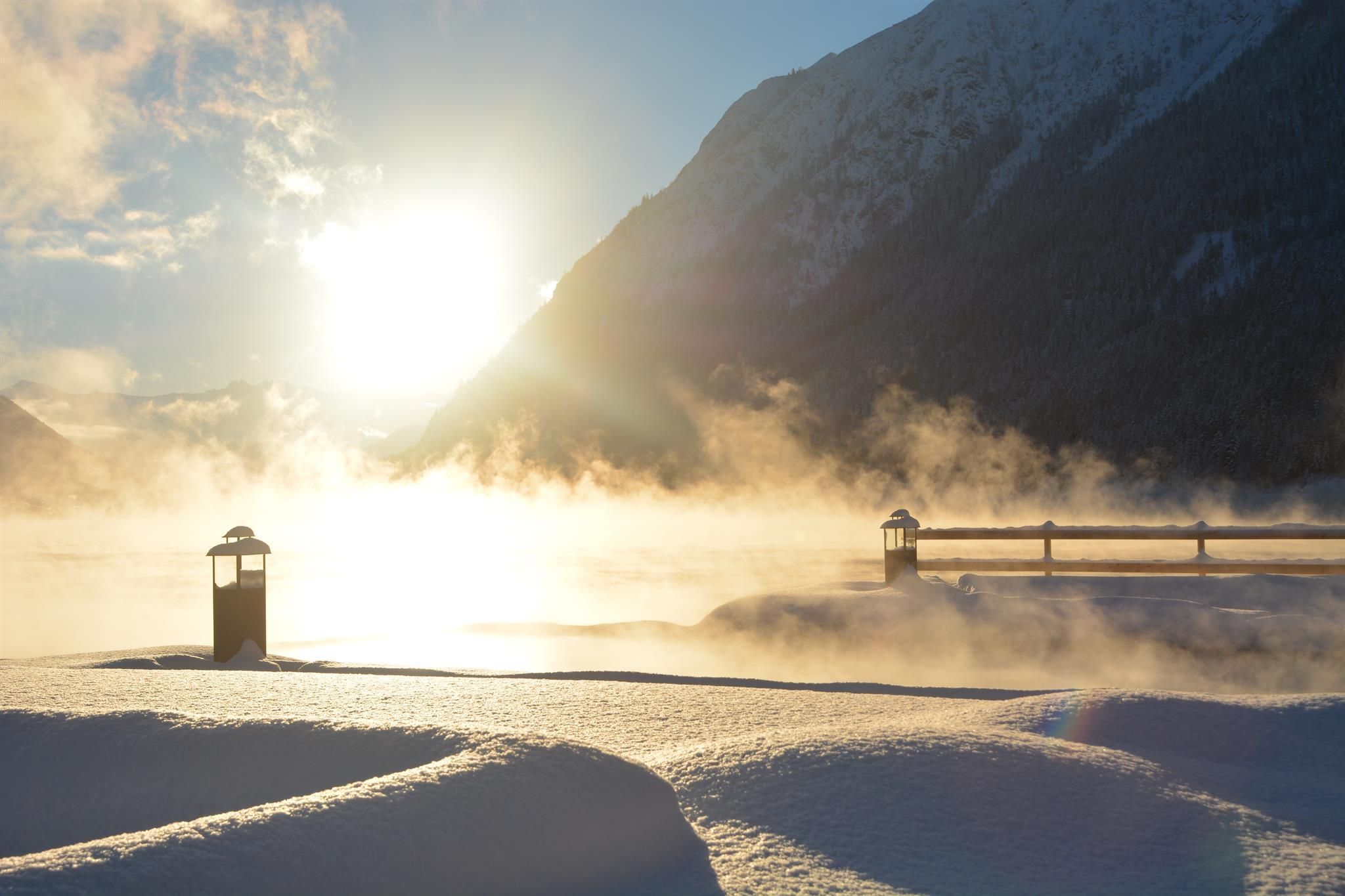 A winter landscape with a snow-covered ground and mountains in the background. The rising sun creates a mystical atmosphere with light fog.