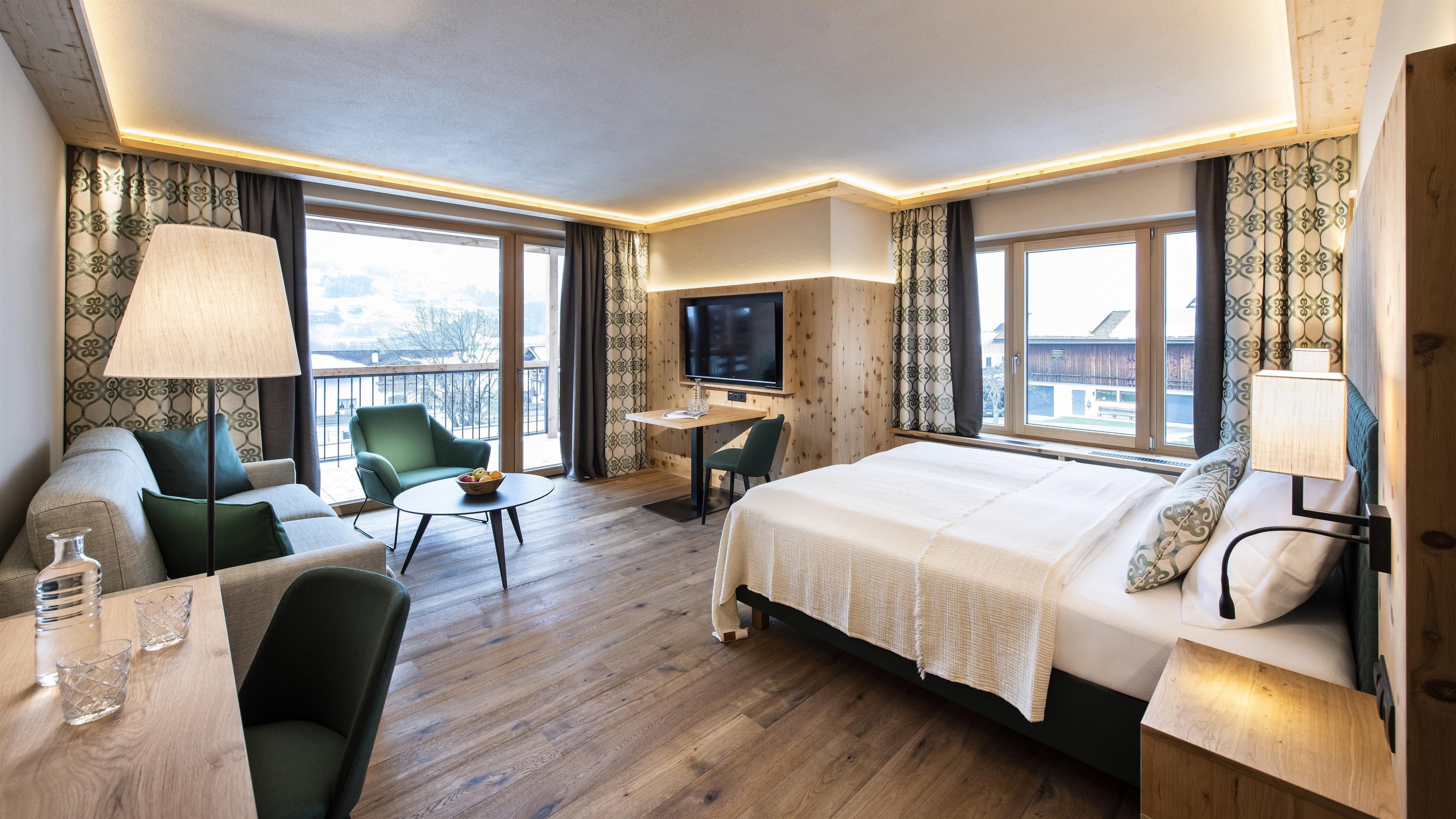 A modern hotel room with a large bed, a cozy living area, and plenty of natural light. The surroundings are stylishly decorated and offer a view of the mountains.