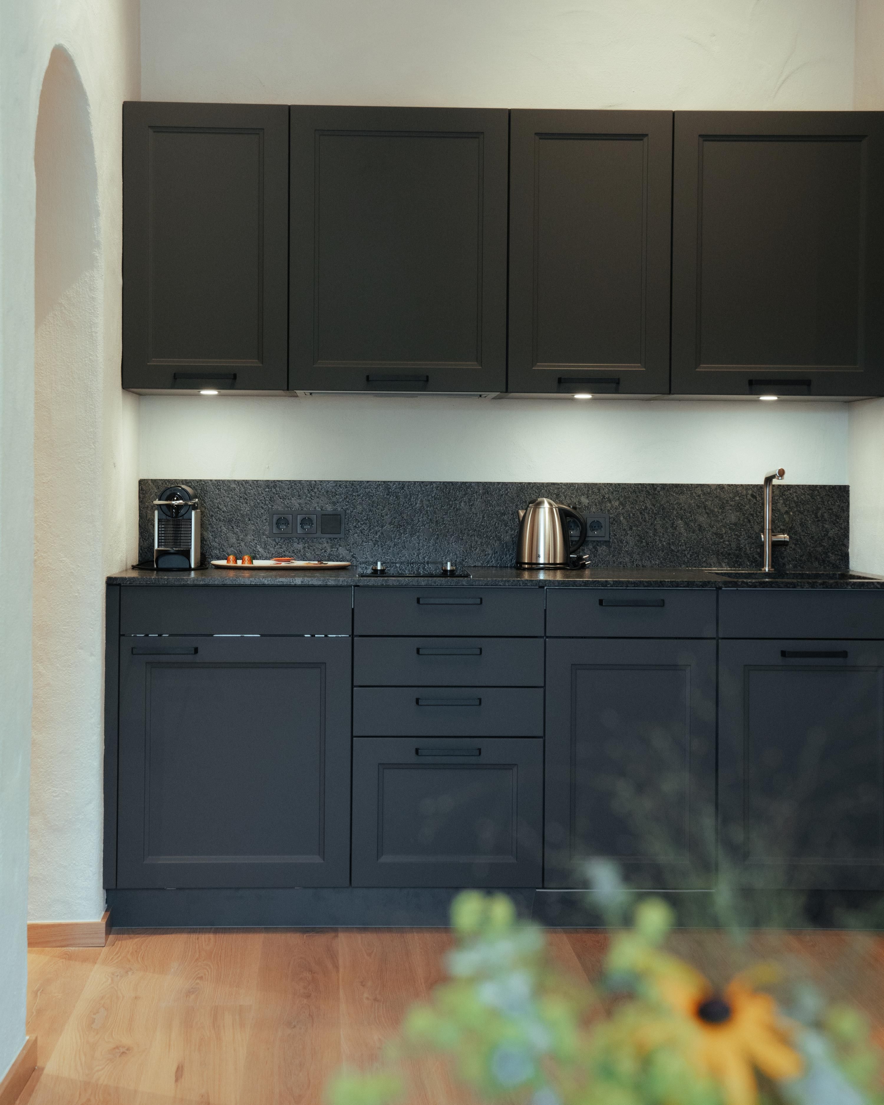 A modern kitchen with black cabinets and an elegant countertop. The equipment includes a coffee maker, a kettle, and a stove.