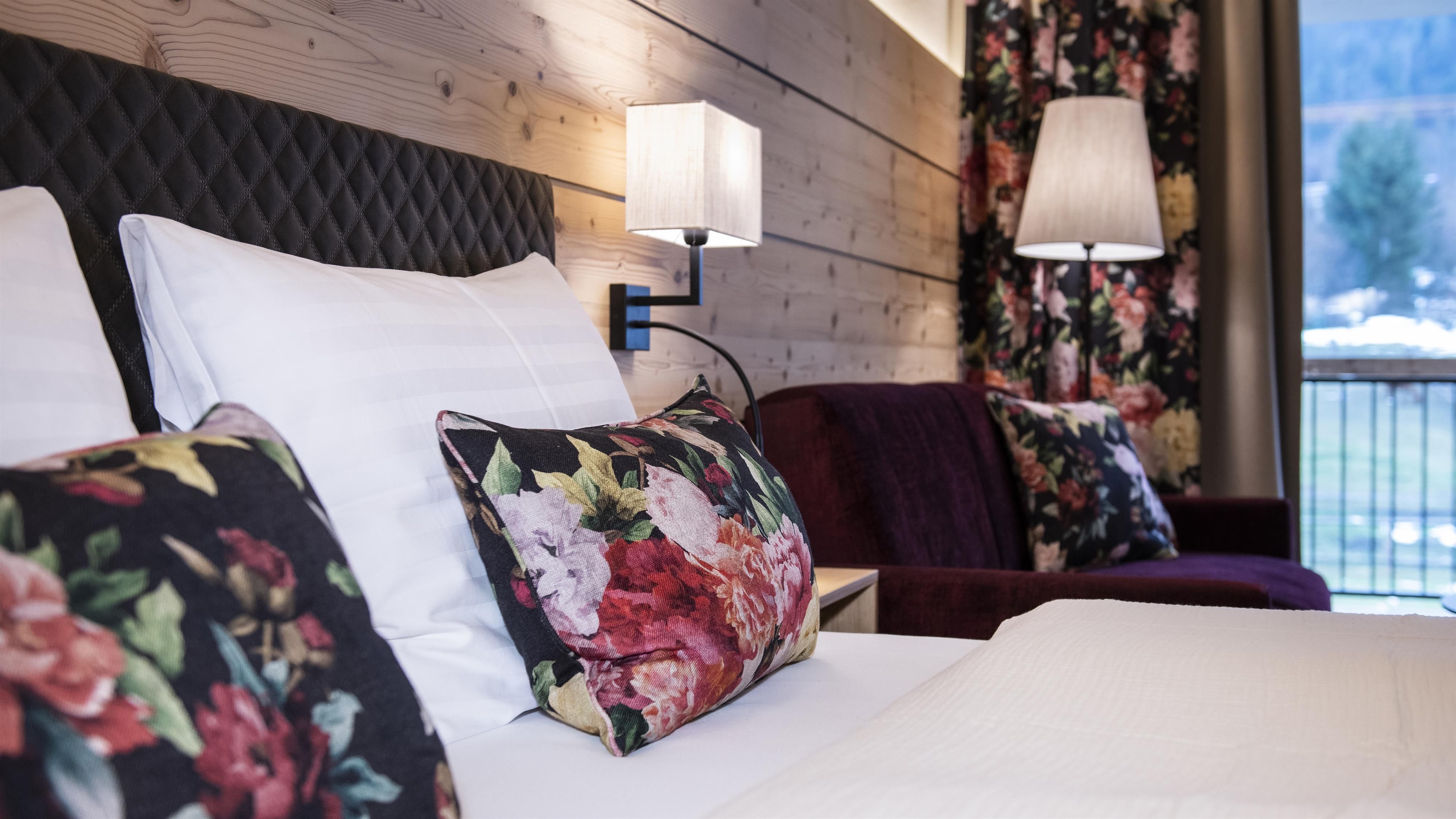 A cozy bedroom with floral patterns on the pillows and a stylish wooden wall. Soft lighting creates an inviting atmosphere.