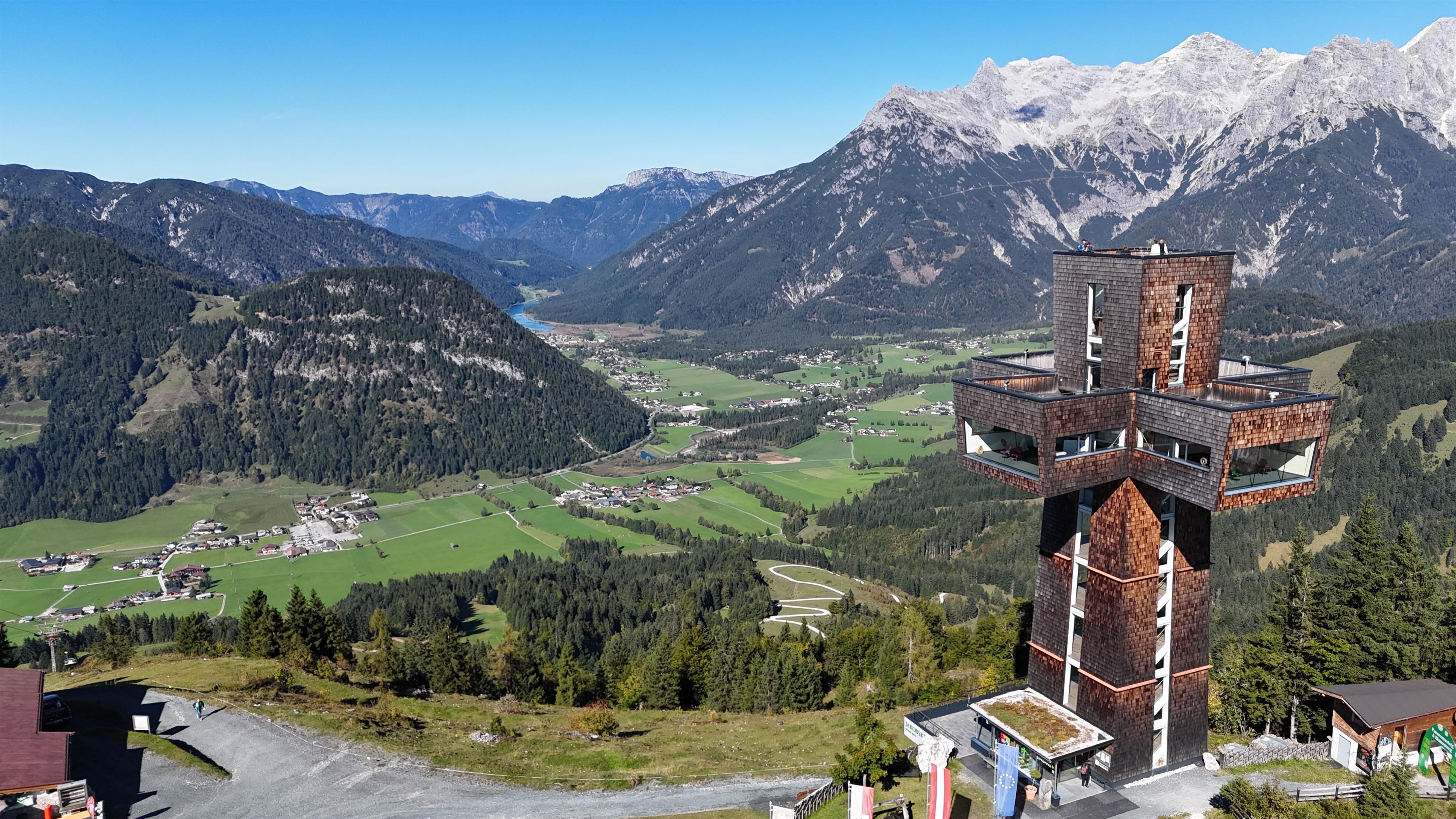 An impressive mountain landscape with a viewing tower in the foreground. In the background, snow-covered mountains and green valleys can be seen.