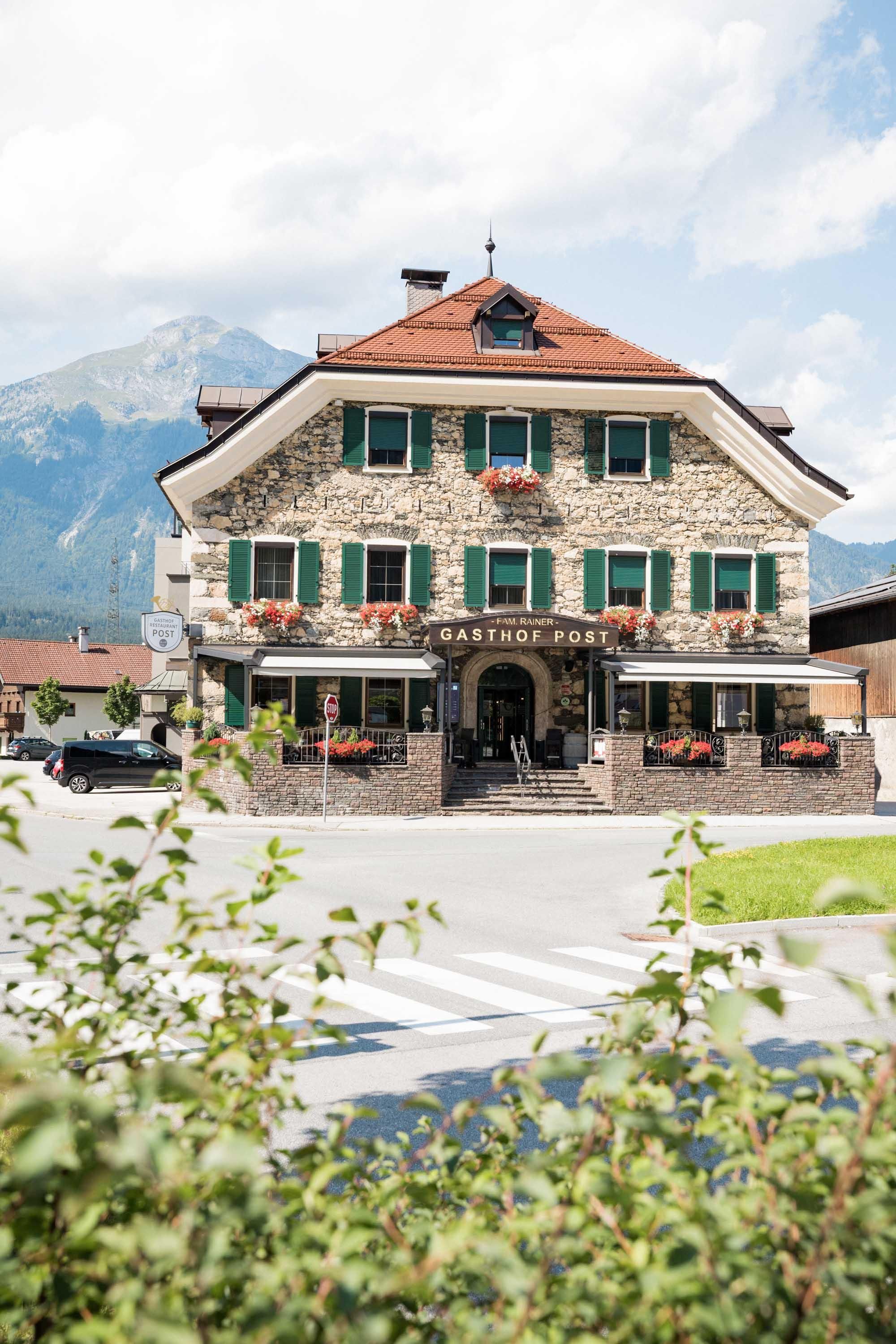A beautiful, traditional inn with green shutters and flowers. In the background, mountains and a blue sky are visible.
