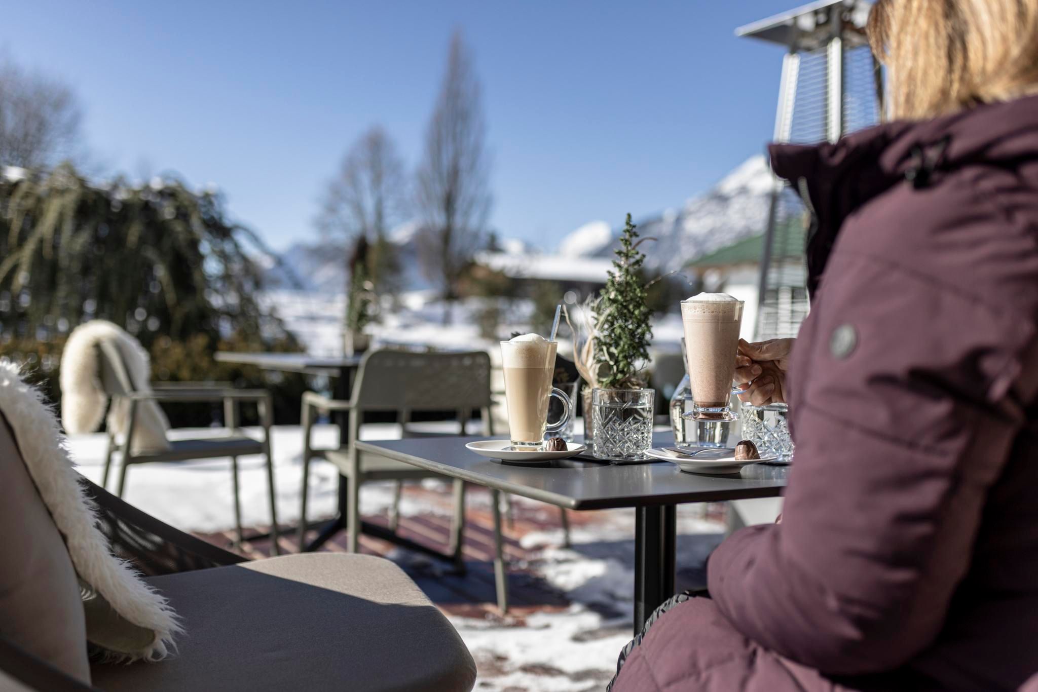 A cozy outdoor area with a table and drinks in the cold season. In the background, snow-covered mountains and a clear blue sky can be seen.