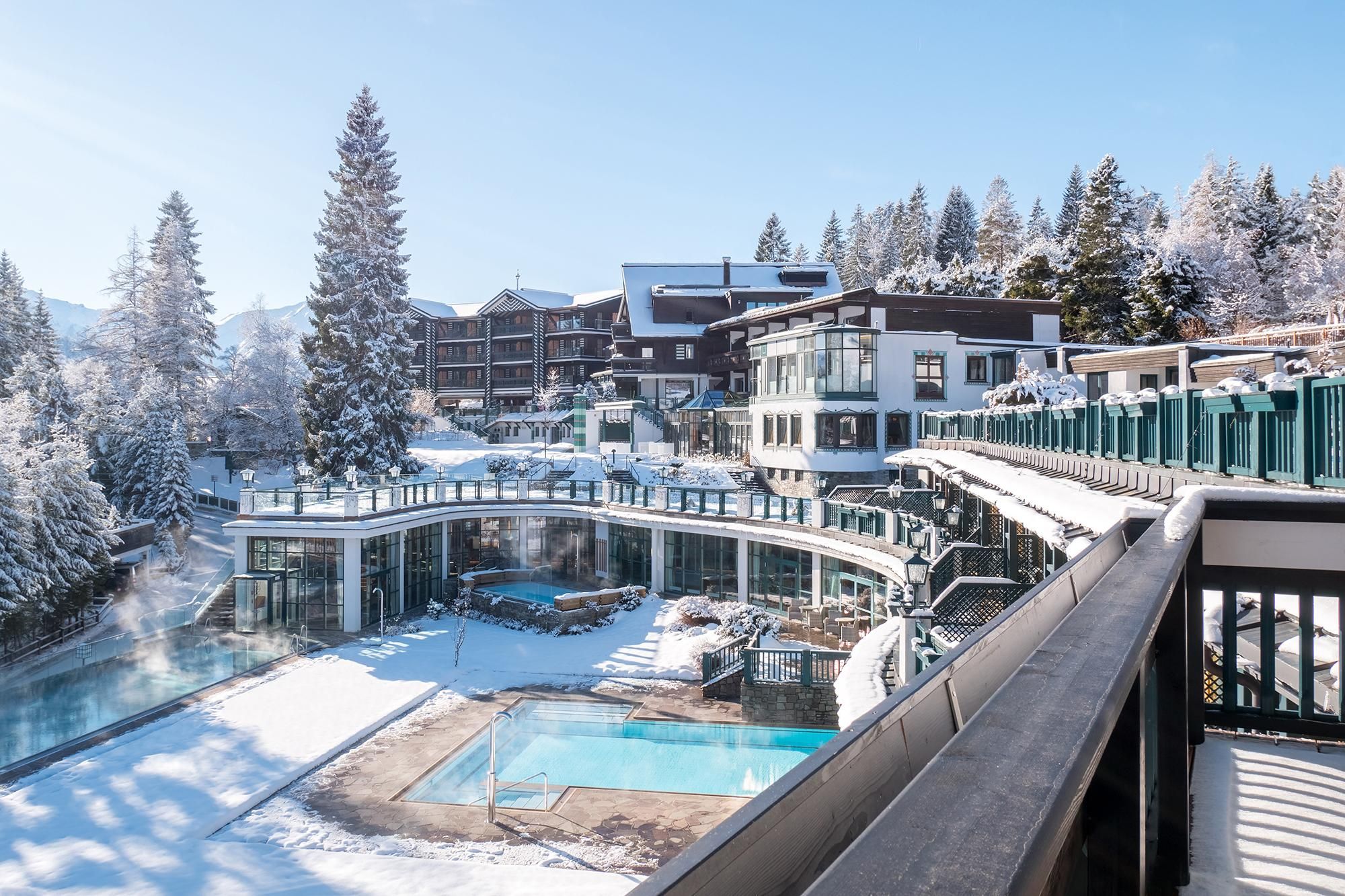 An elegant hotel in a snowy landscape. In the foreground, there are outdoor pools and modernly designed terraces.