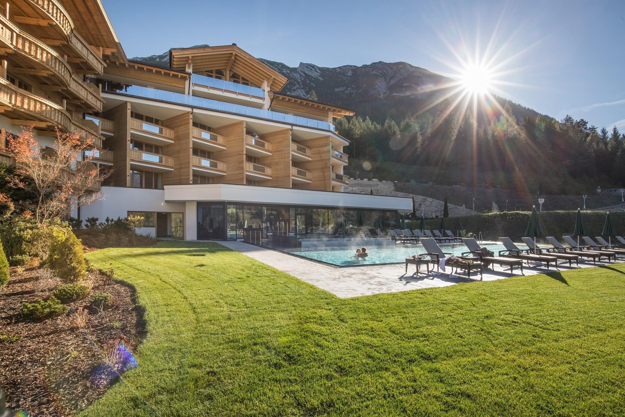 A modern hotel with a pool and loungers in the garden. The sun is shining over the mountain landscape.