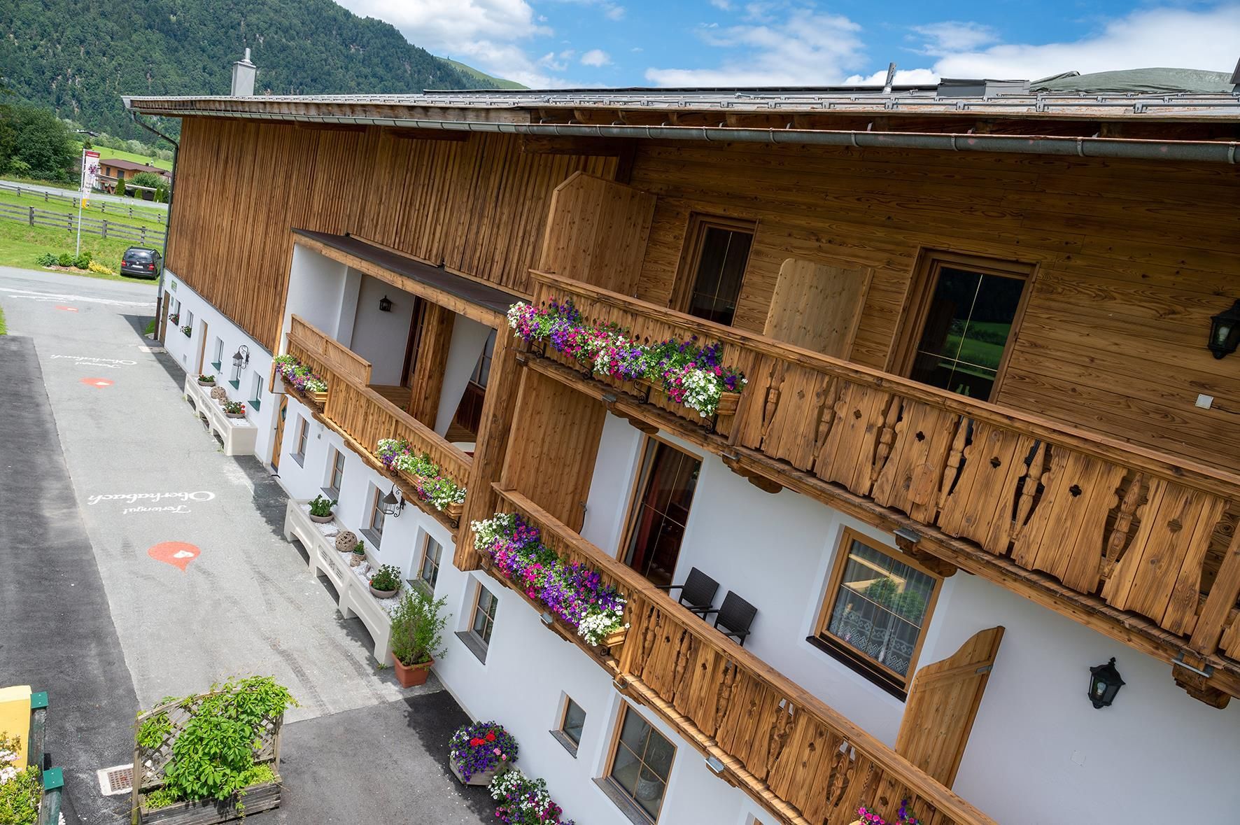 A wooden building with balconies decorated with colorful flowers. In the background, green mountains and a blue sky can be seen.
