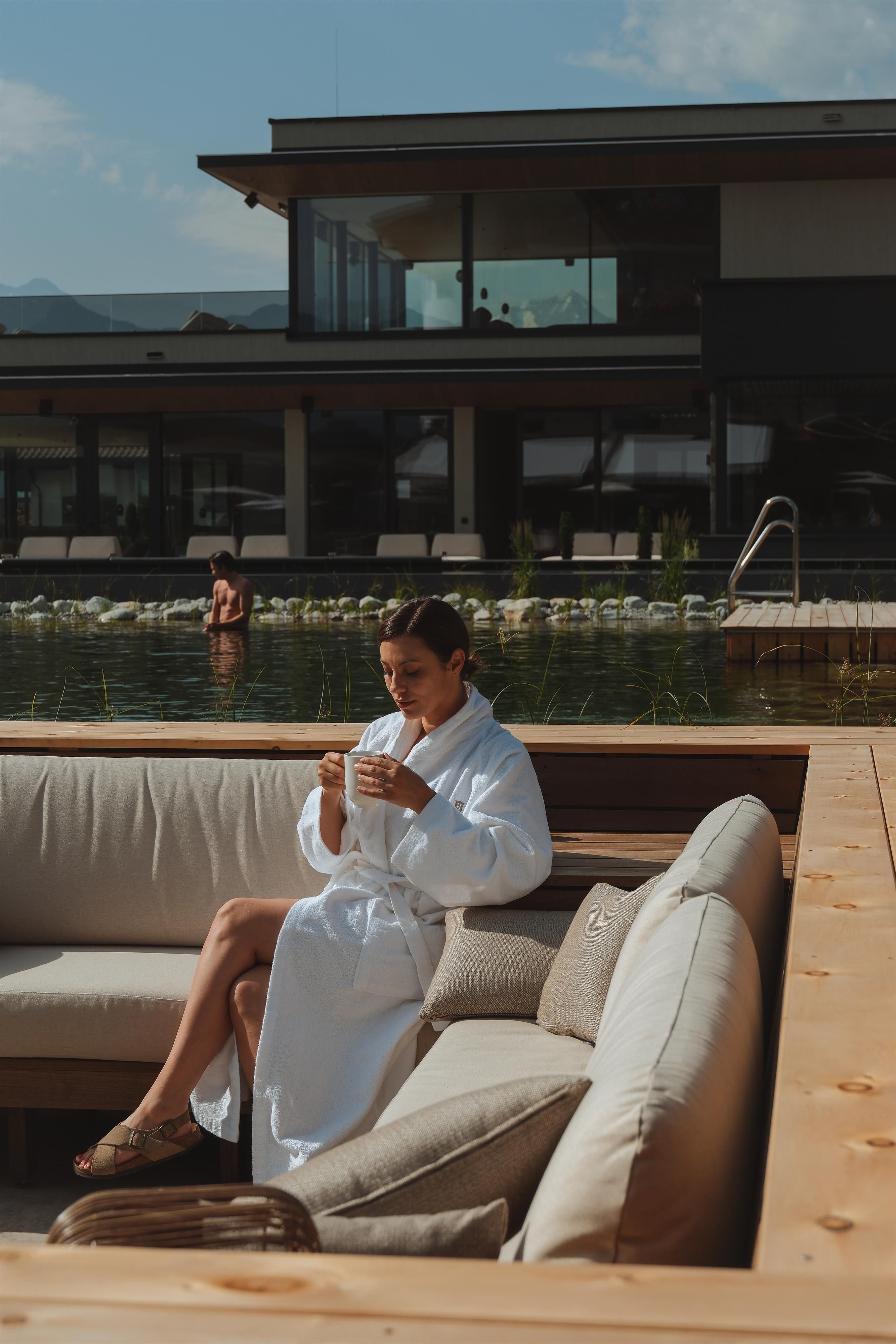 A woman in a bathrobe sits relaxed on a sofa landscape and holds a cup. In the background, a pool and a modern building are visible.