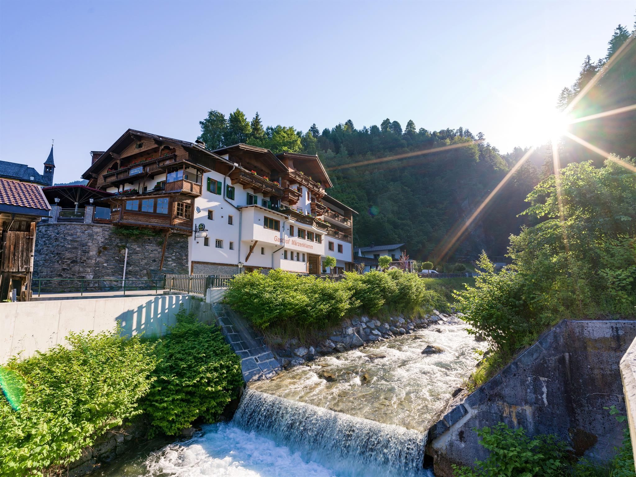 A picturesque building on the banks of a rushing stream. In the background, the sun shines over the green mountains.